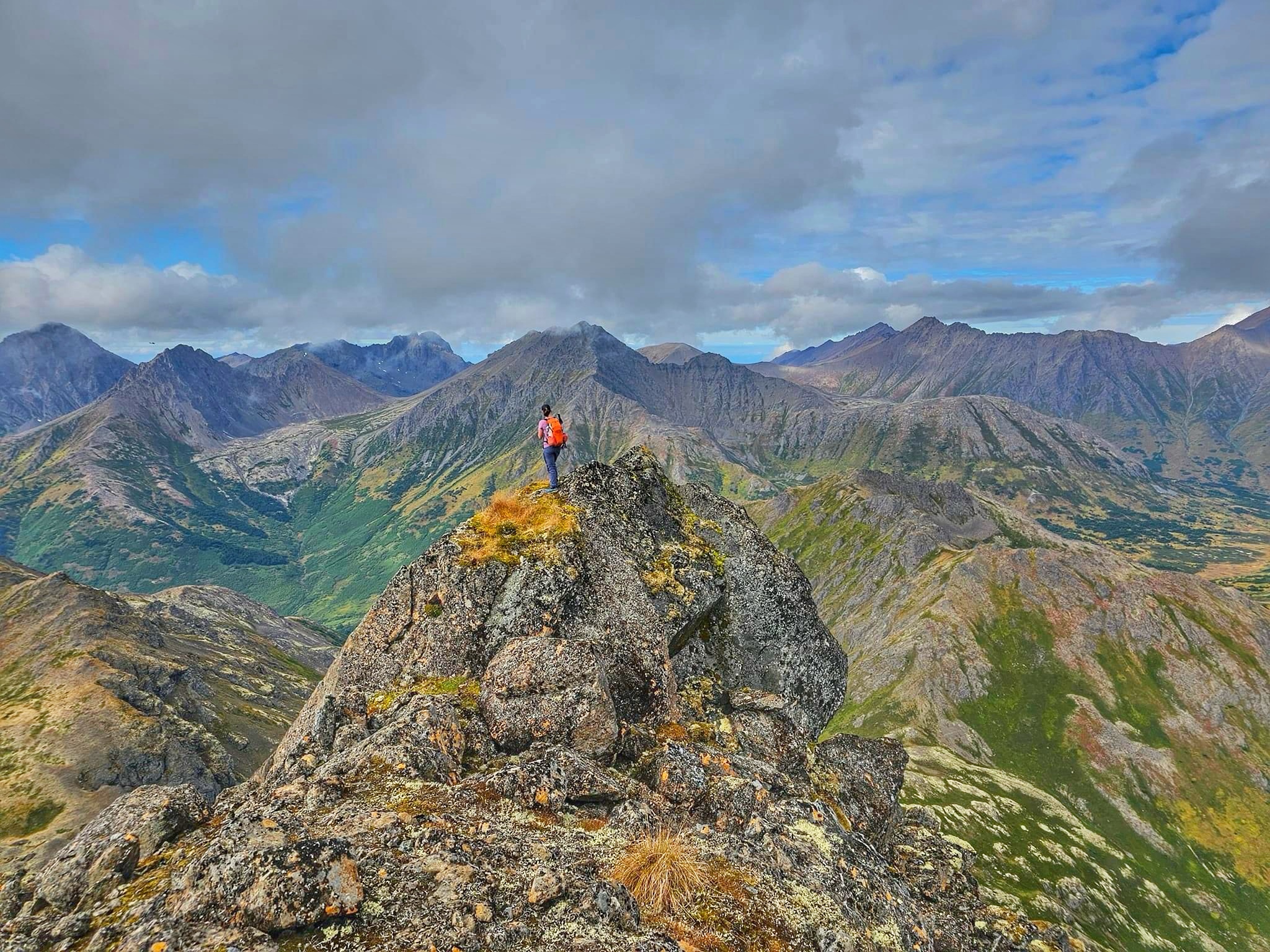 A person stands on top of a mountain peak looking at the mountains in the distance.  The sky is partly cloudy, partly clear and the vegetation is bright with the fall colors. 