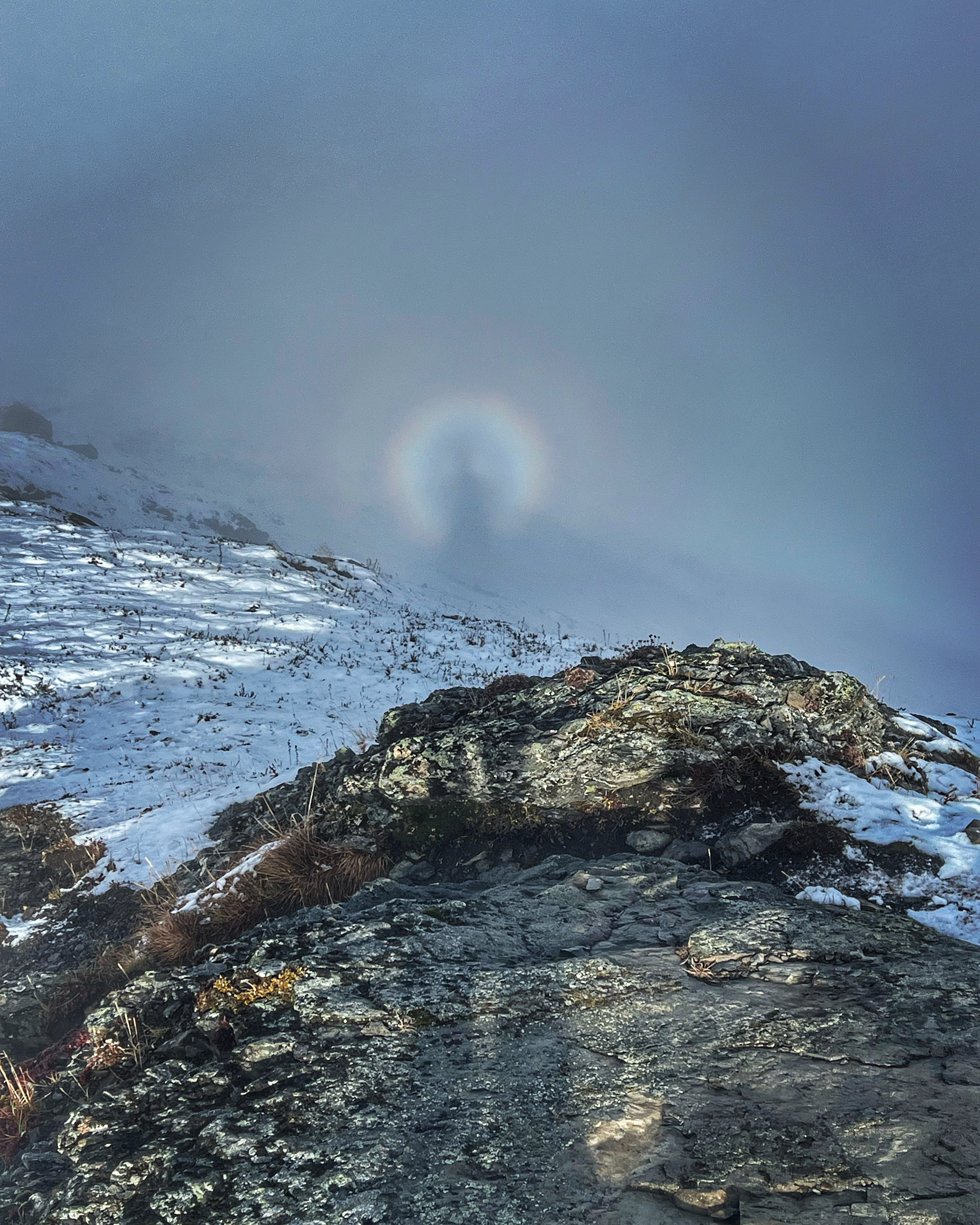 A person's elongated shadow extends over rocks and onto the slope of a mountain. A rainbow halo surrounds the upper portion of the shadow