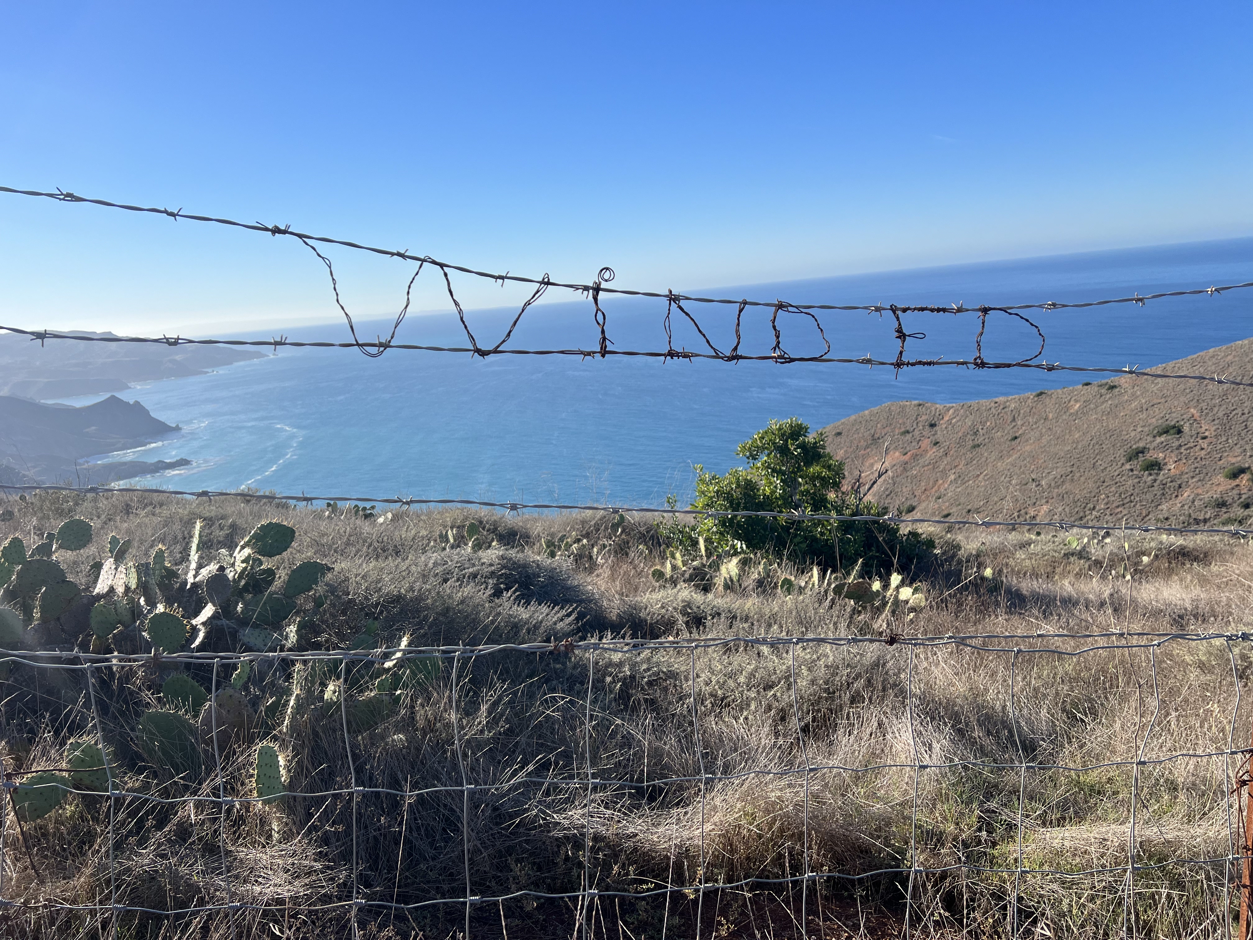 Wire wrapped around barbed wire fence to spell out "winded" overlooking ocean