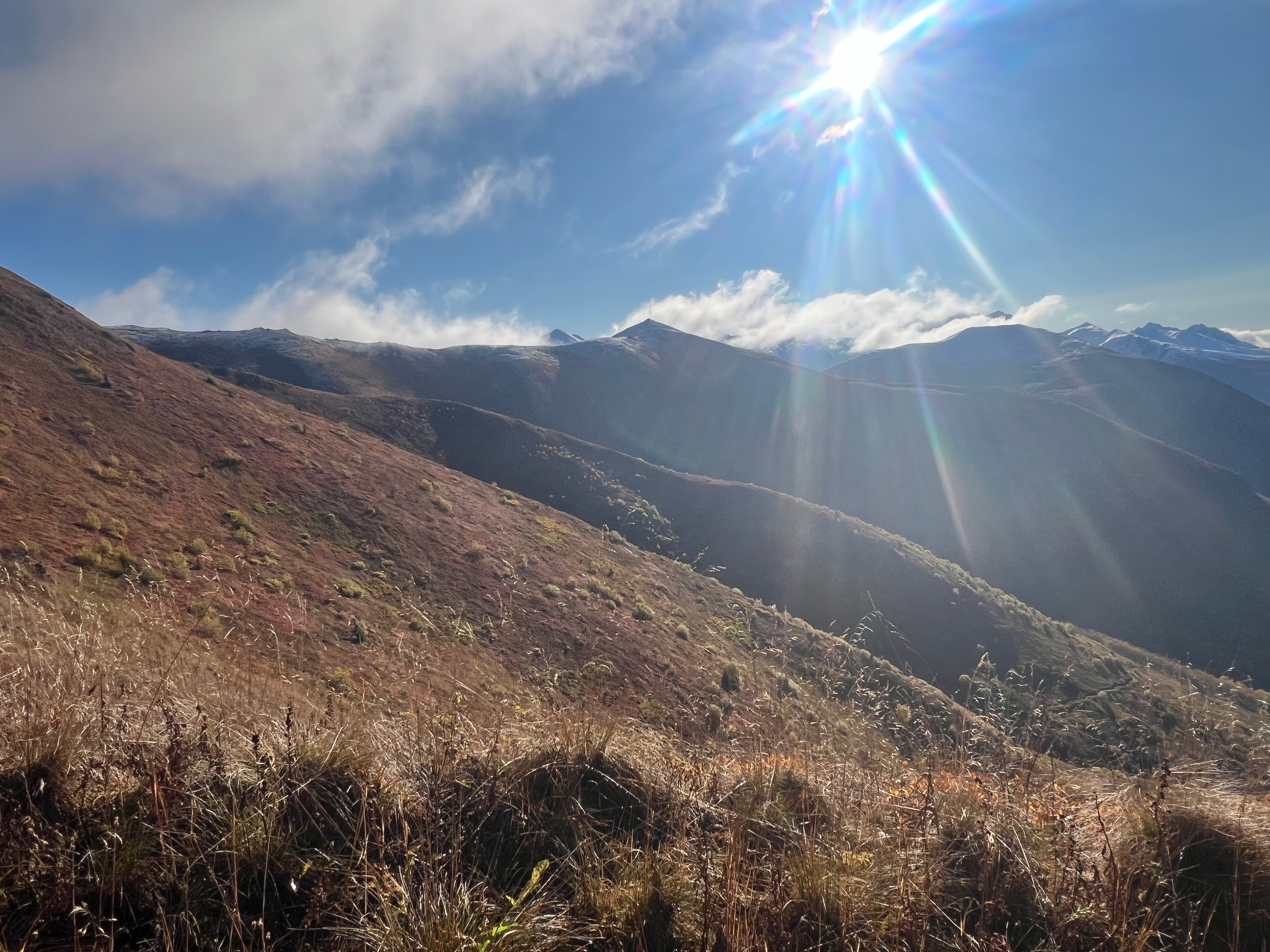 The sun shines above while clouds cap some of the mountains. A snow dusted ridge ends in a pointy peak
