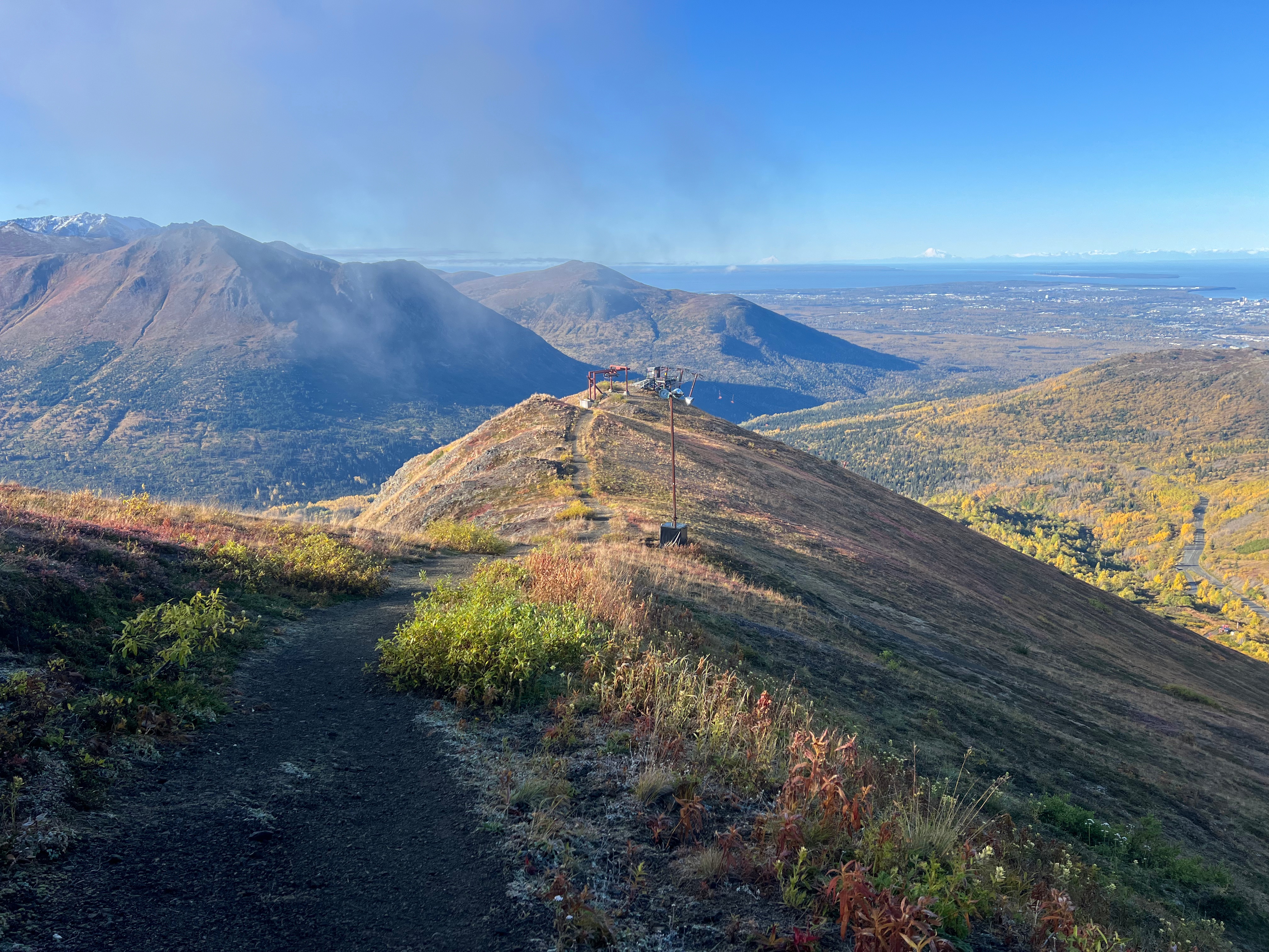 Facing down the ridgeline, a wide dirt path leads down to the top of a ski lift. Mountains and the Cook inlet visible in the distance