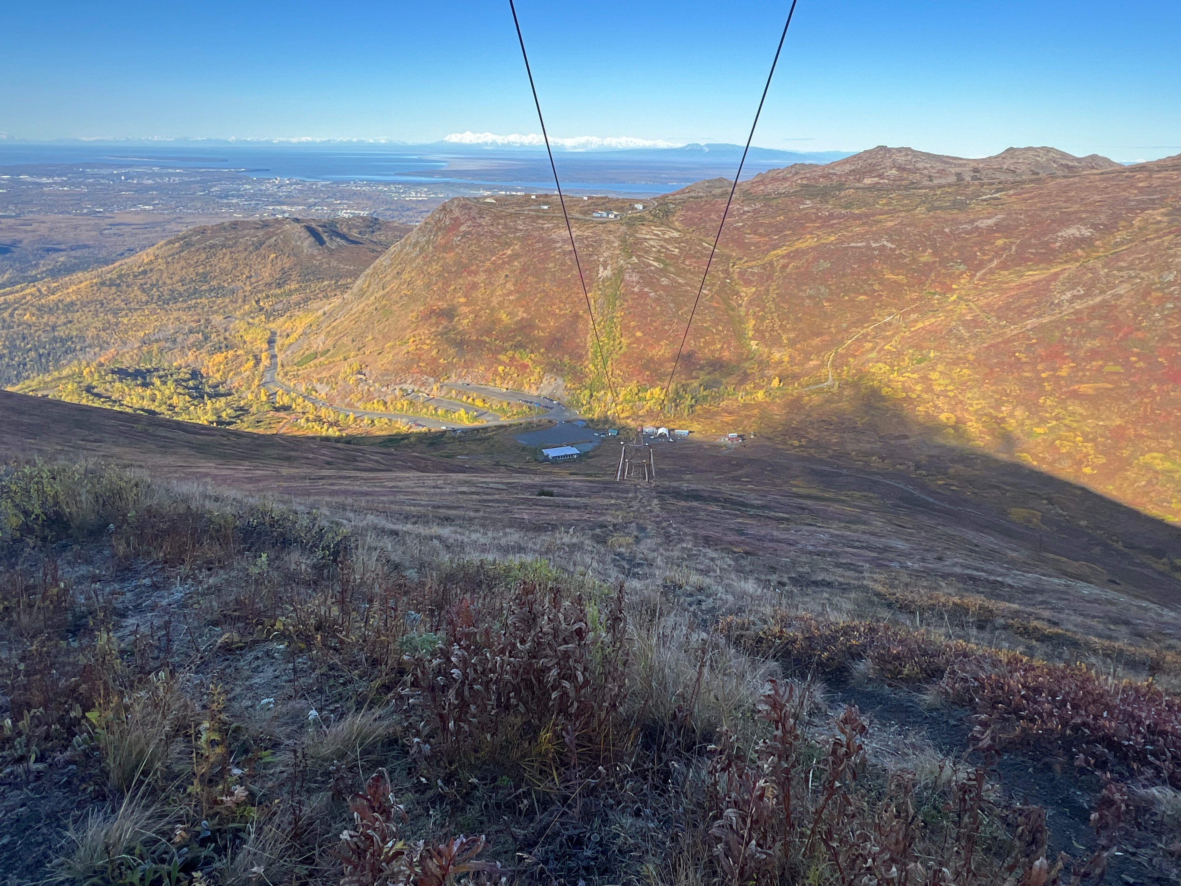Facing downhill, two cables run up from the bottom. T-bar ski lift infrastructure hold up the parallel cables. A parking lot and buildings are at the bottom of the hill.