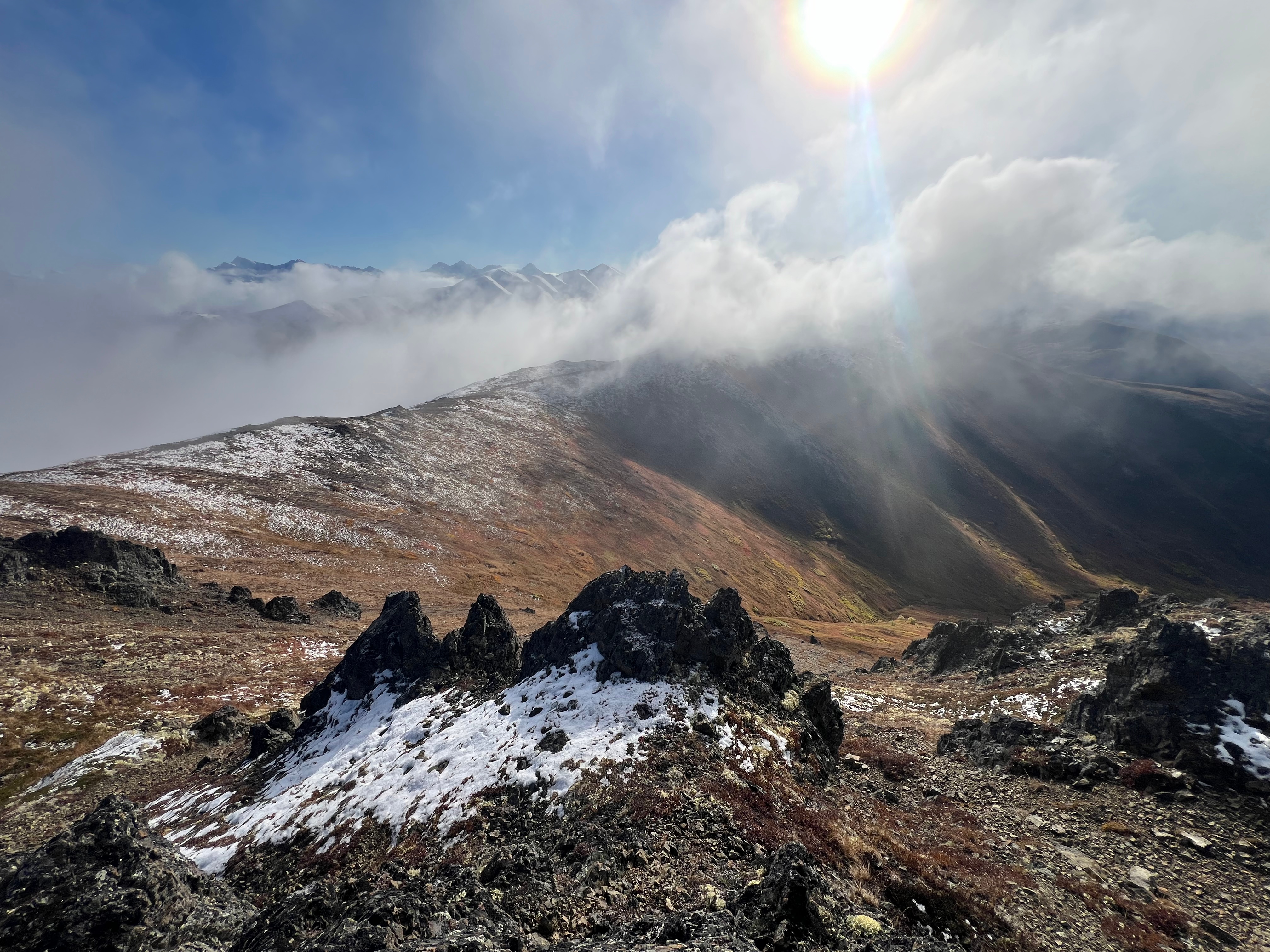 The sun shines brightly above a cloud of fog covering peak 3891. The ridge is dusted in snow. Many mountains are in the distance. Pointed, sharp rocks in the foreground