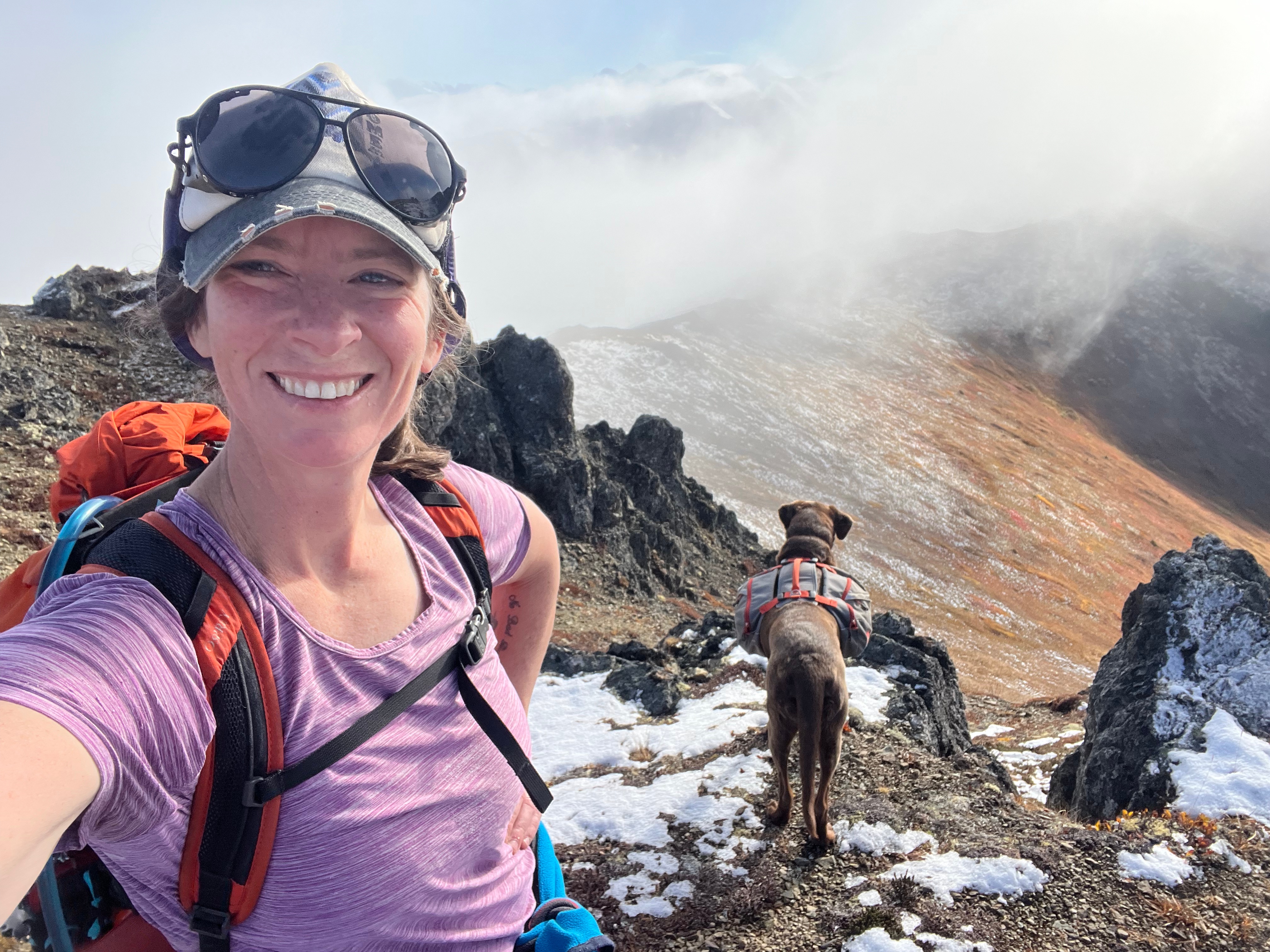 The author taking a selfie with her dog looking towards the cloud covered ridgeline.