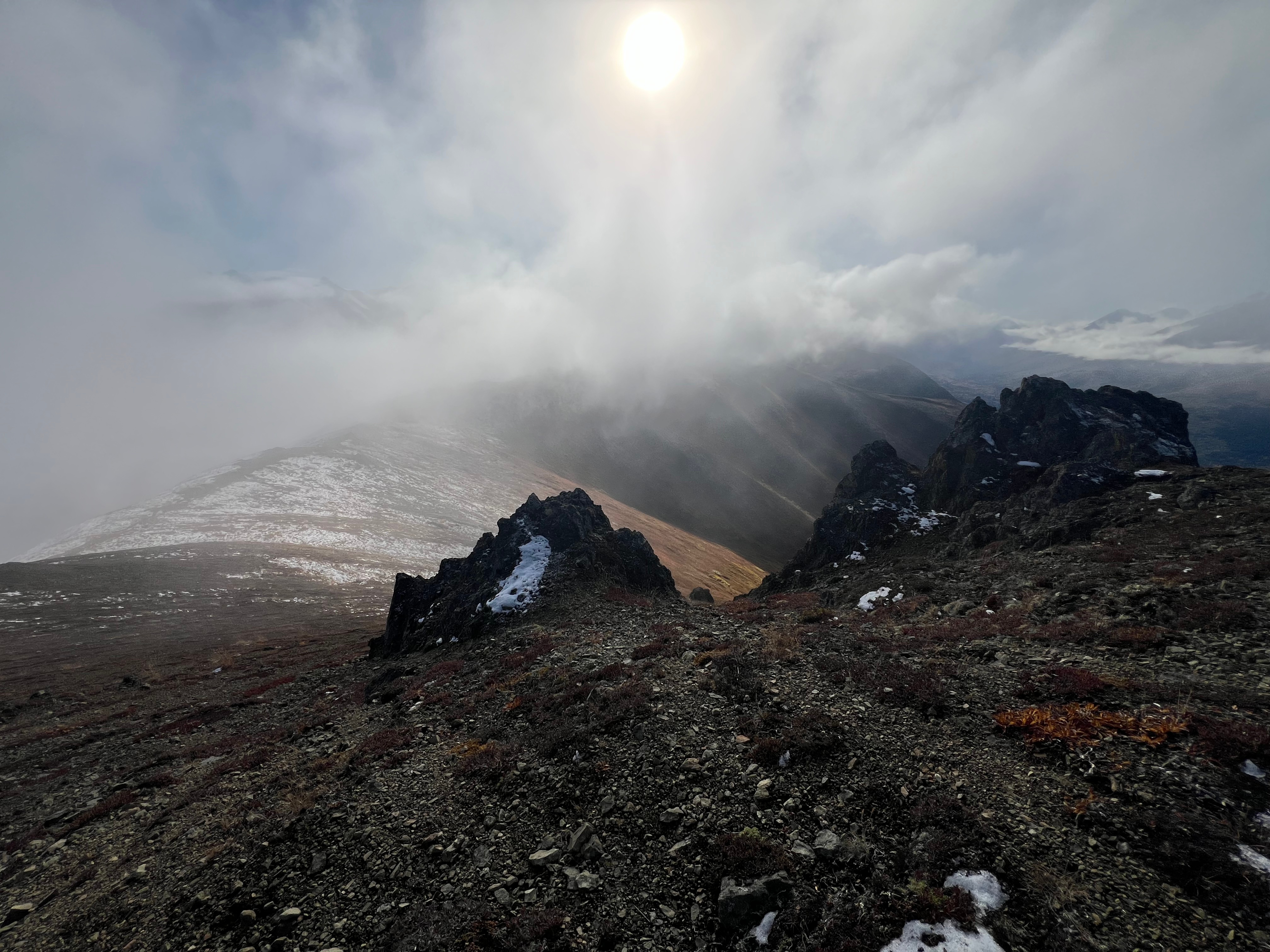 The sun shines brightly above a cloud of fog covering peak 3891. The ridge is dusted in snow. Many mountains are in the distance. Pointed, sharp rocks in the foreground