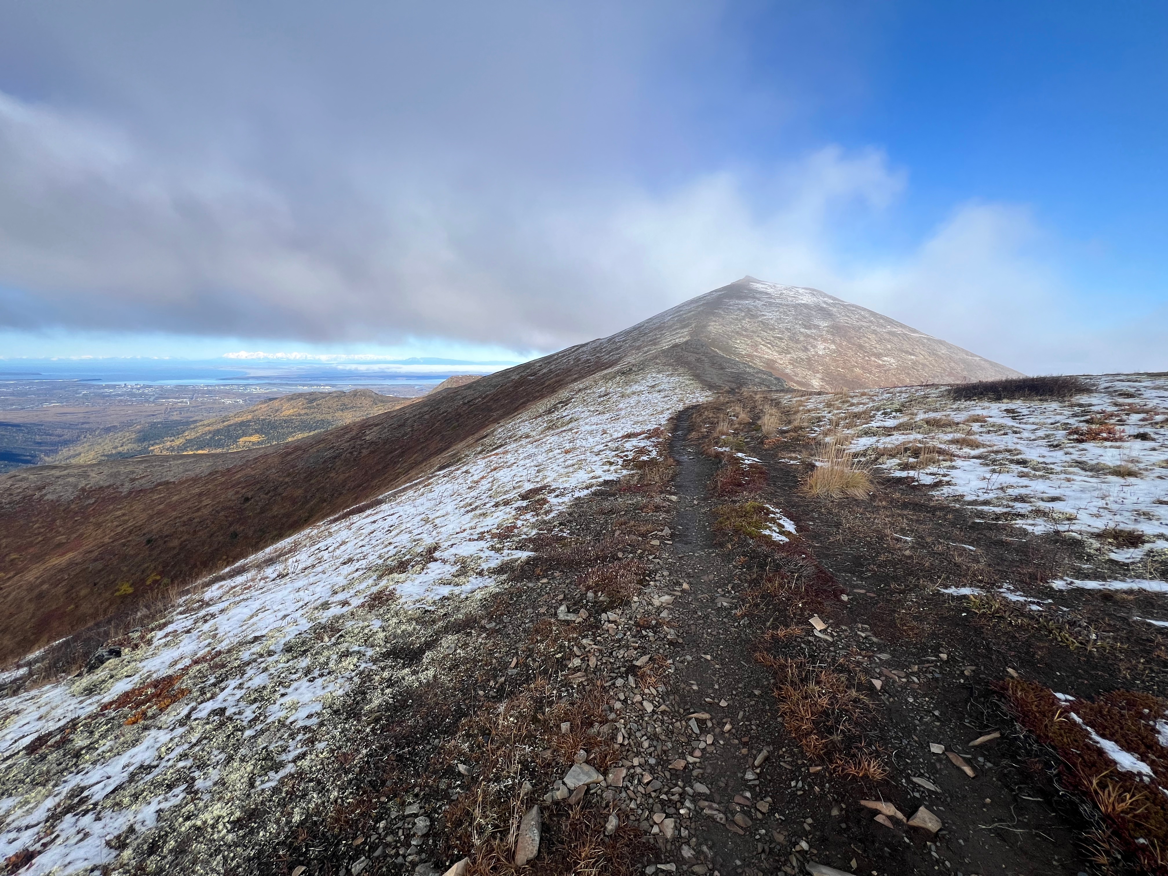 The pointy peak of 3891 is above. A dirt path leading to it is bordered by a dusting of snow. Blue sky is above the light fog.