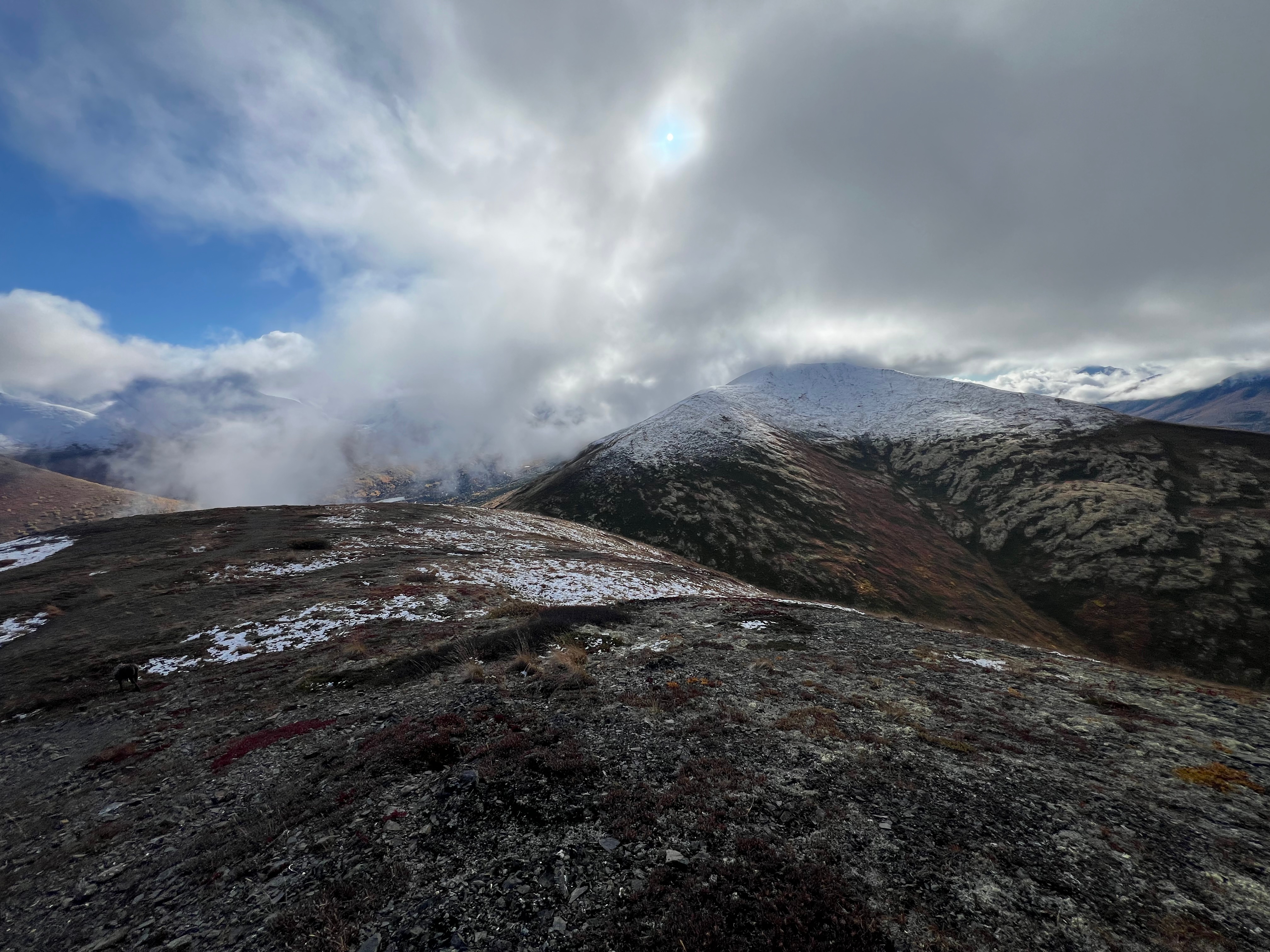 Clouds cover the sun, while blue skies are in the distance. A broad, snowcapped peak on the other side of the low saddle.