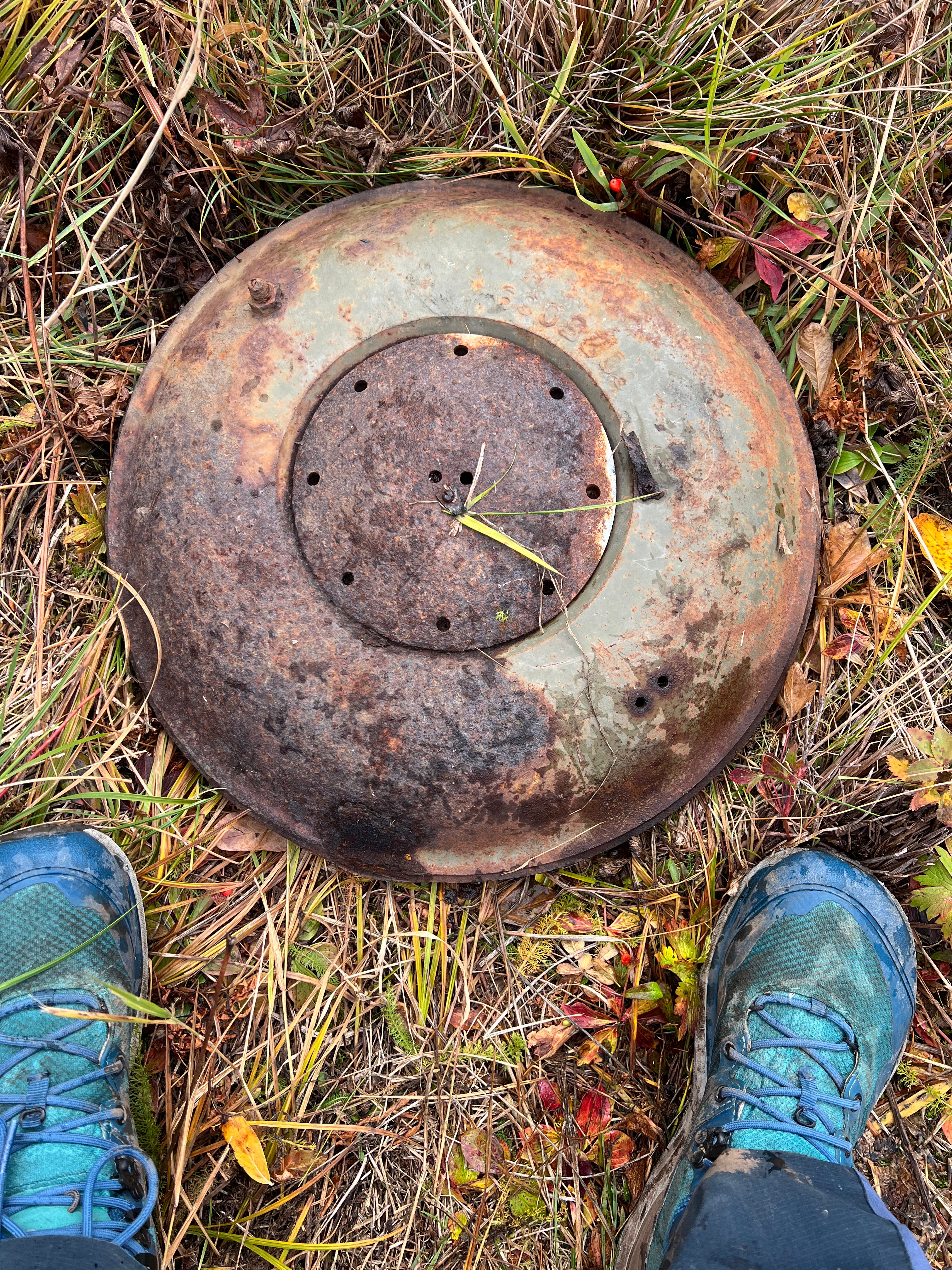 A round metal object lays on the ground. Feet in photos for scale. The rusty piece of missile is about 1' in diameter