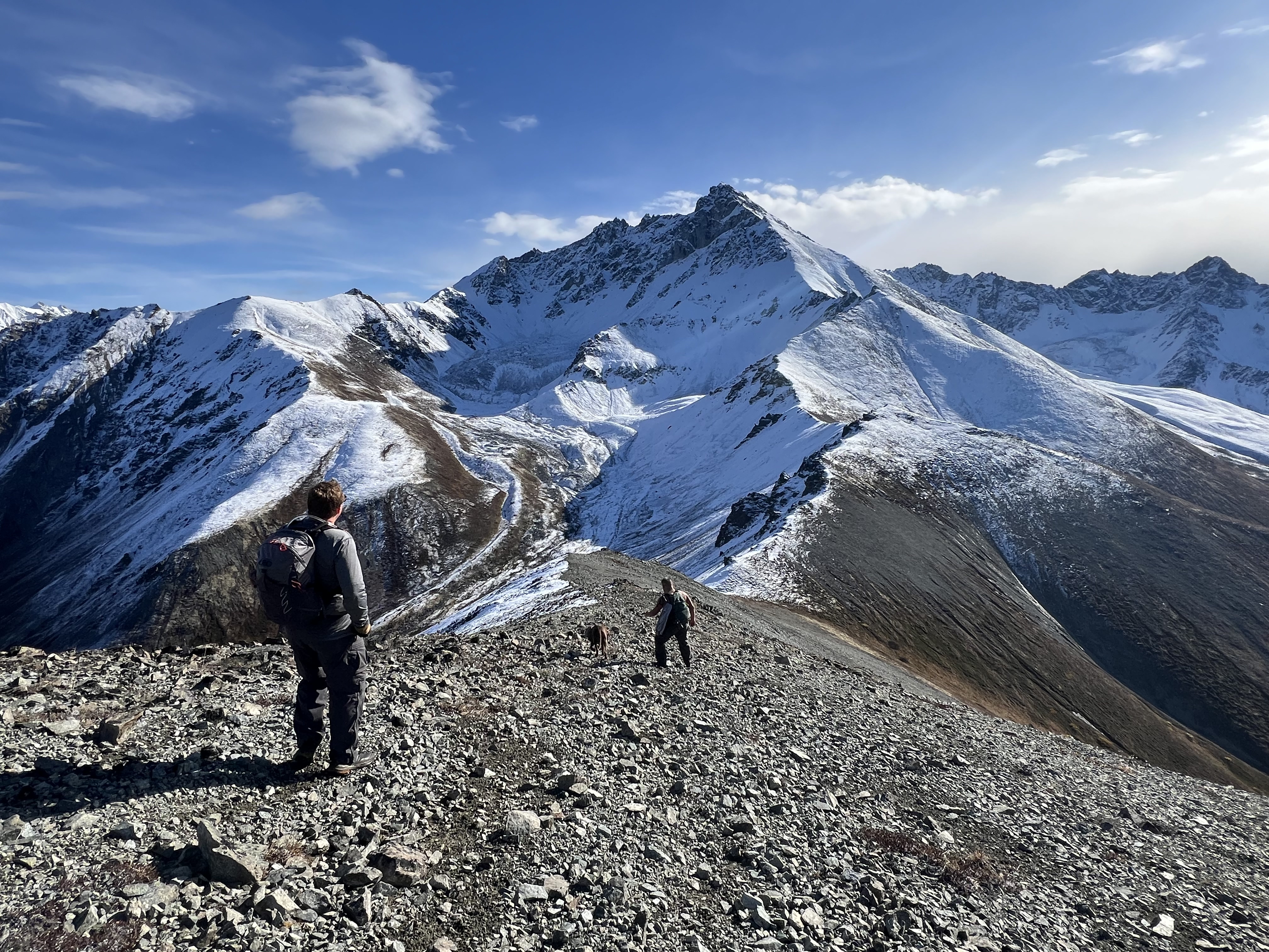 Friends and dog walking down a slope on the ridgeline with giant, snowcapped Mat Peak beyond