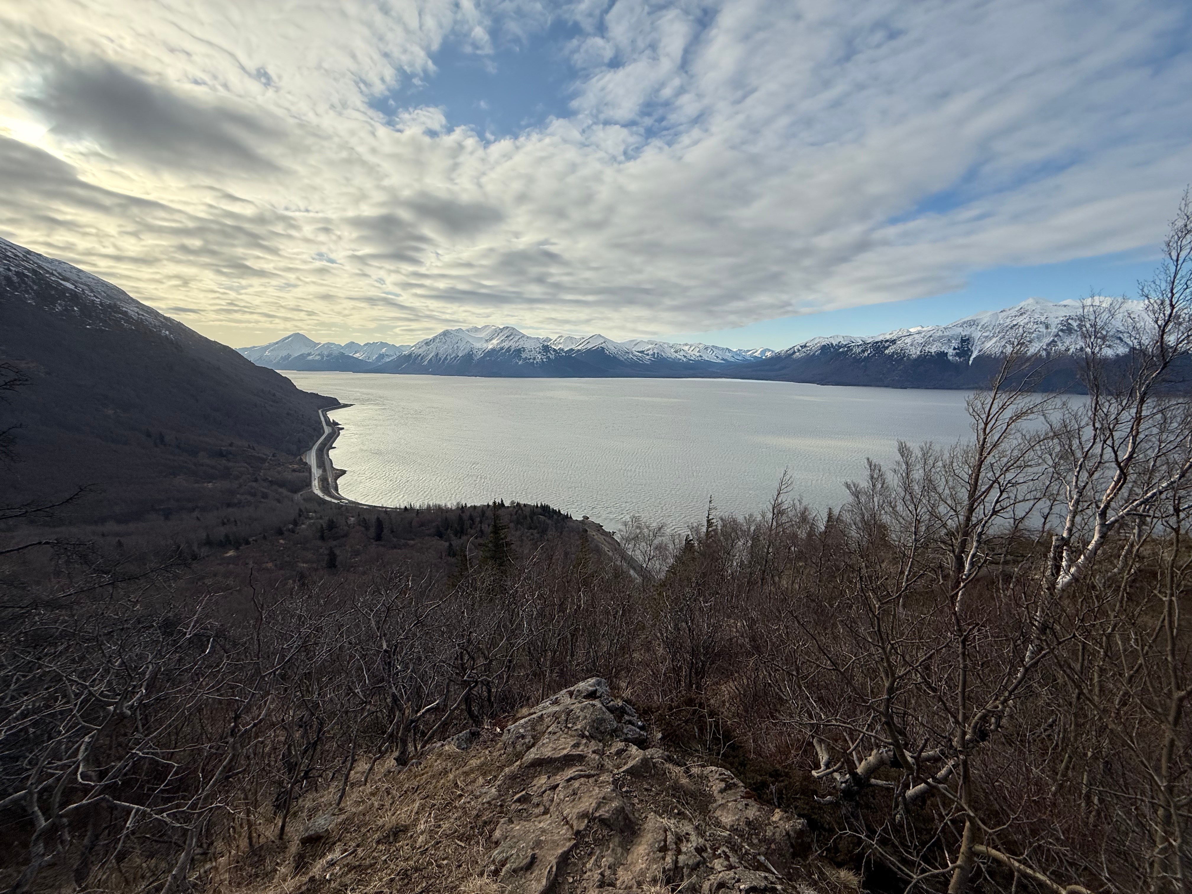 Clouds and blue sky mix above the Turnagain Arm ocean water. The highway is below and snowcapped mountains rise up across the ocean.