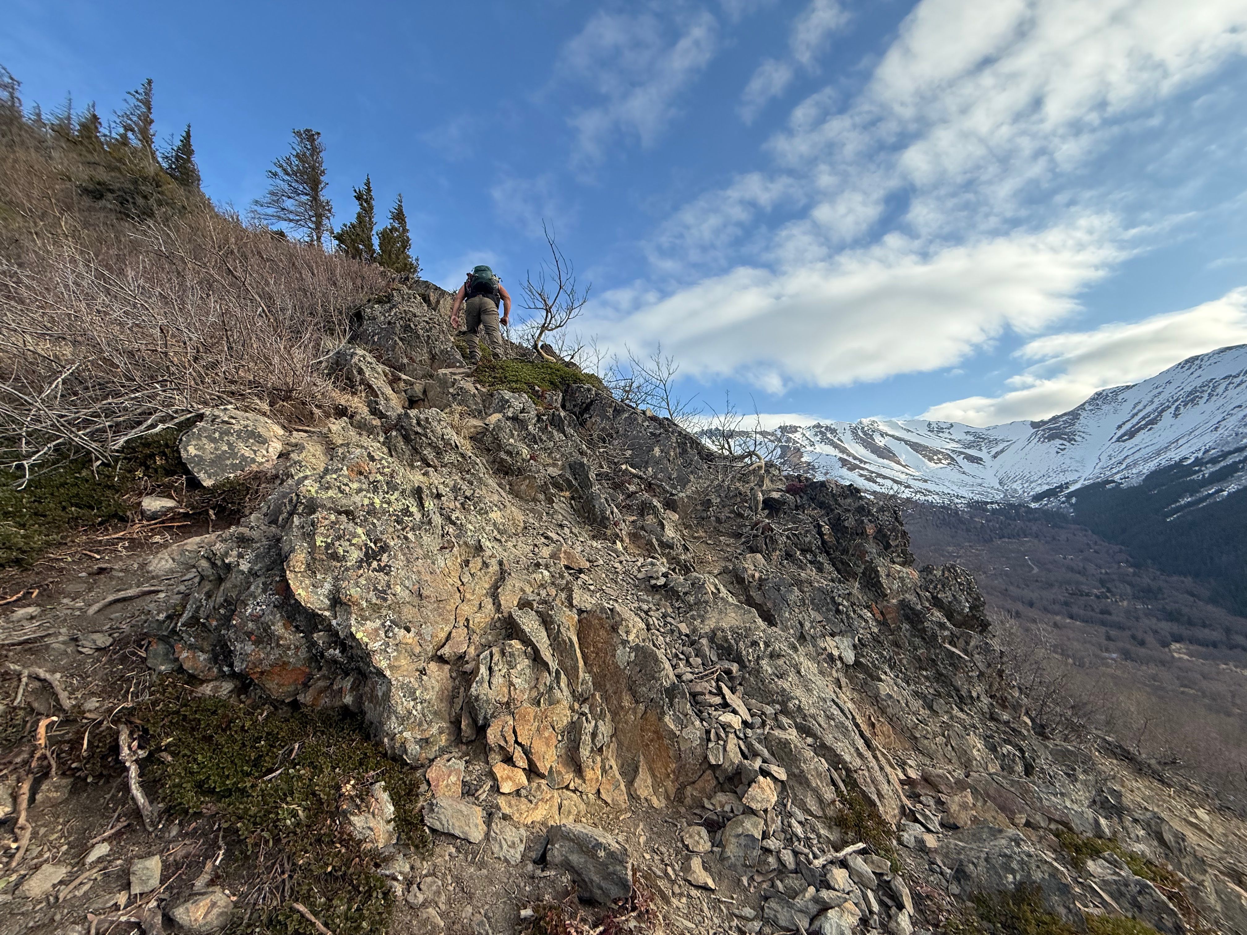 A hiker negotiates a steep, rocky path. Snowcapped mountains rise up at the end of the valley