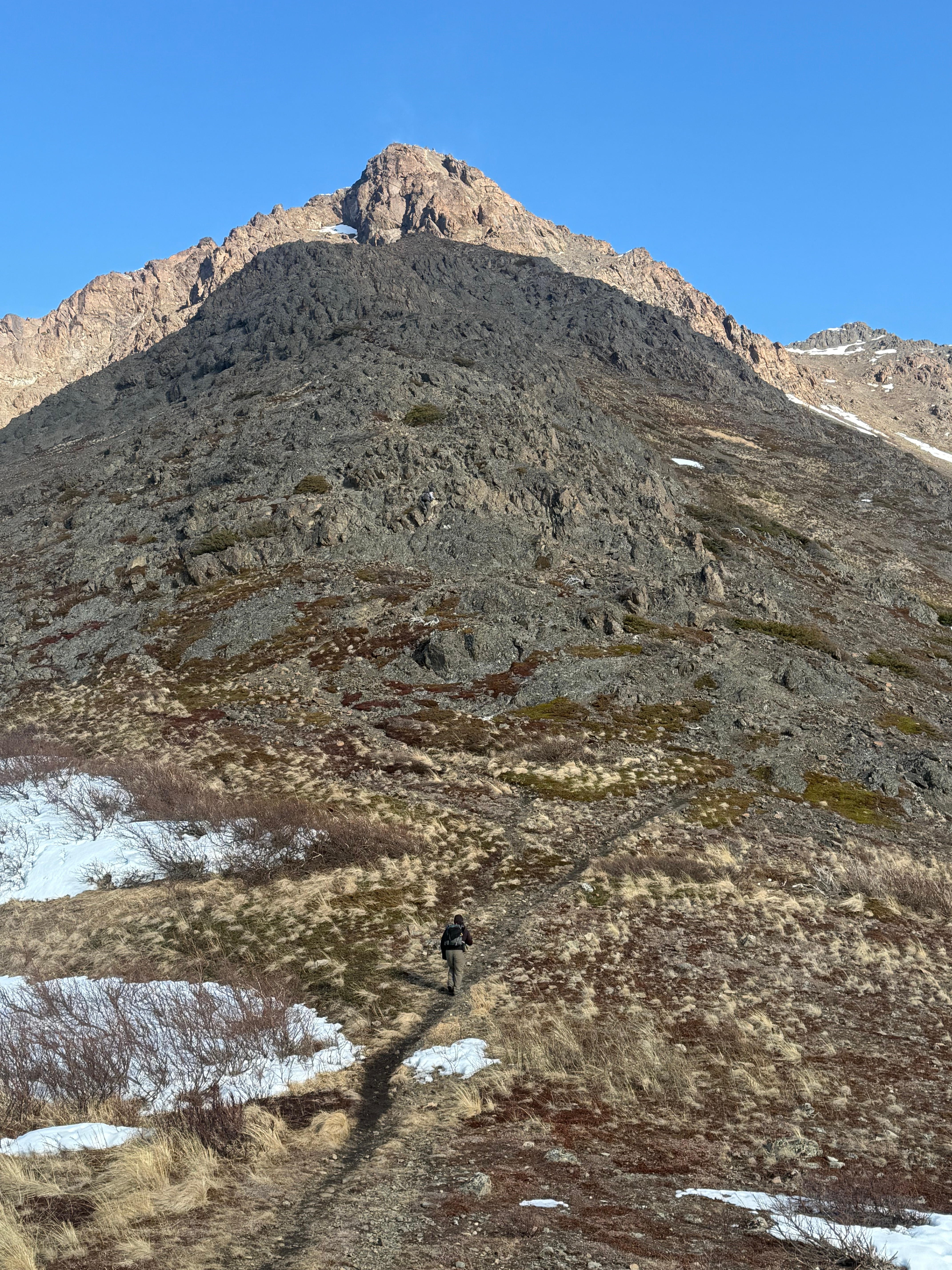 A hiker walks up a narrow dirt footpath. The summit is above. Vegetation dots the slopes.