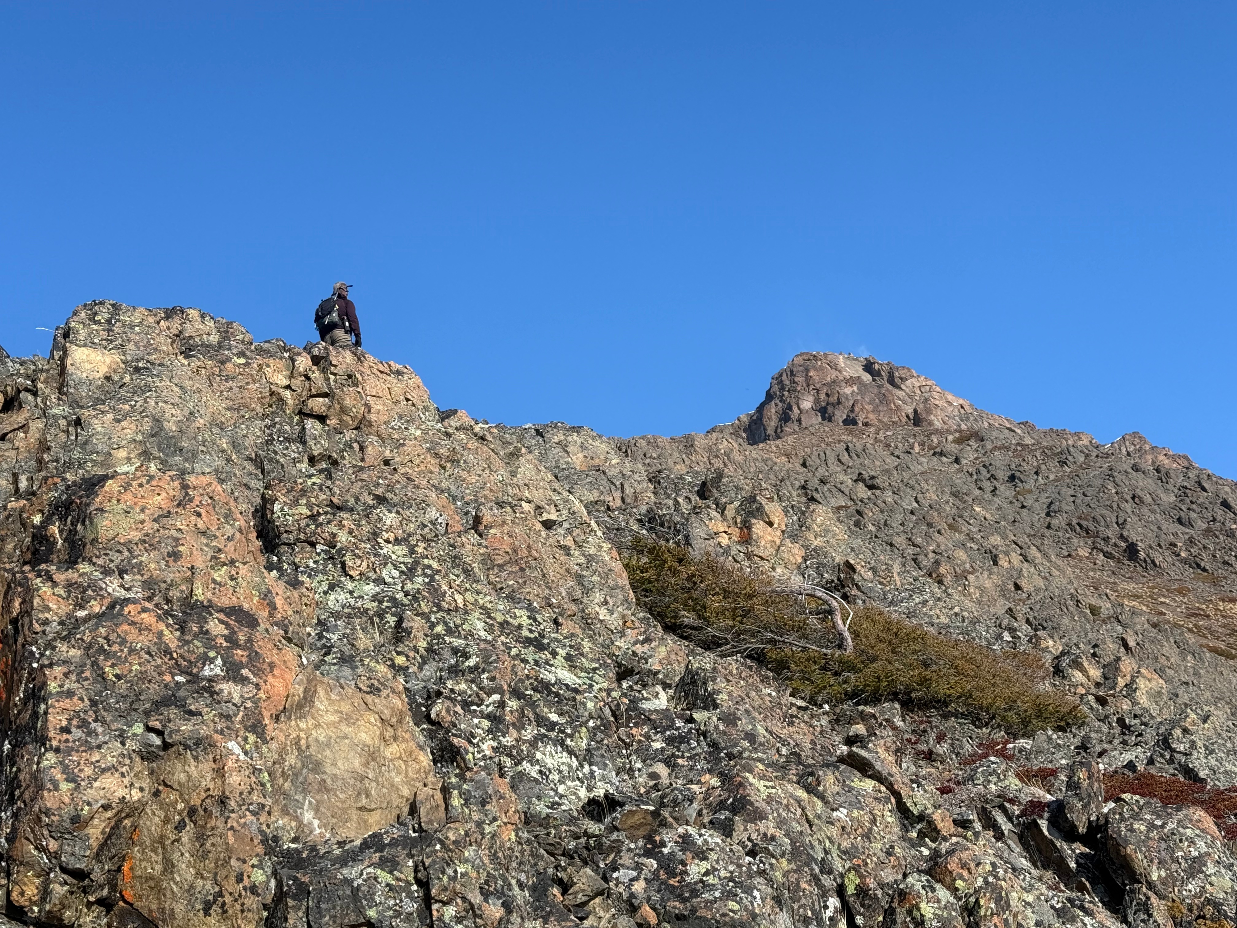 A hiker stand high on a rock looking up at the summit. Snow is blowing off of the peak.