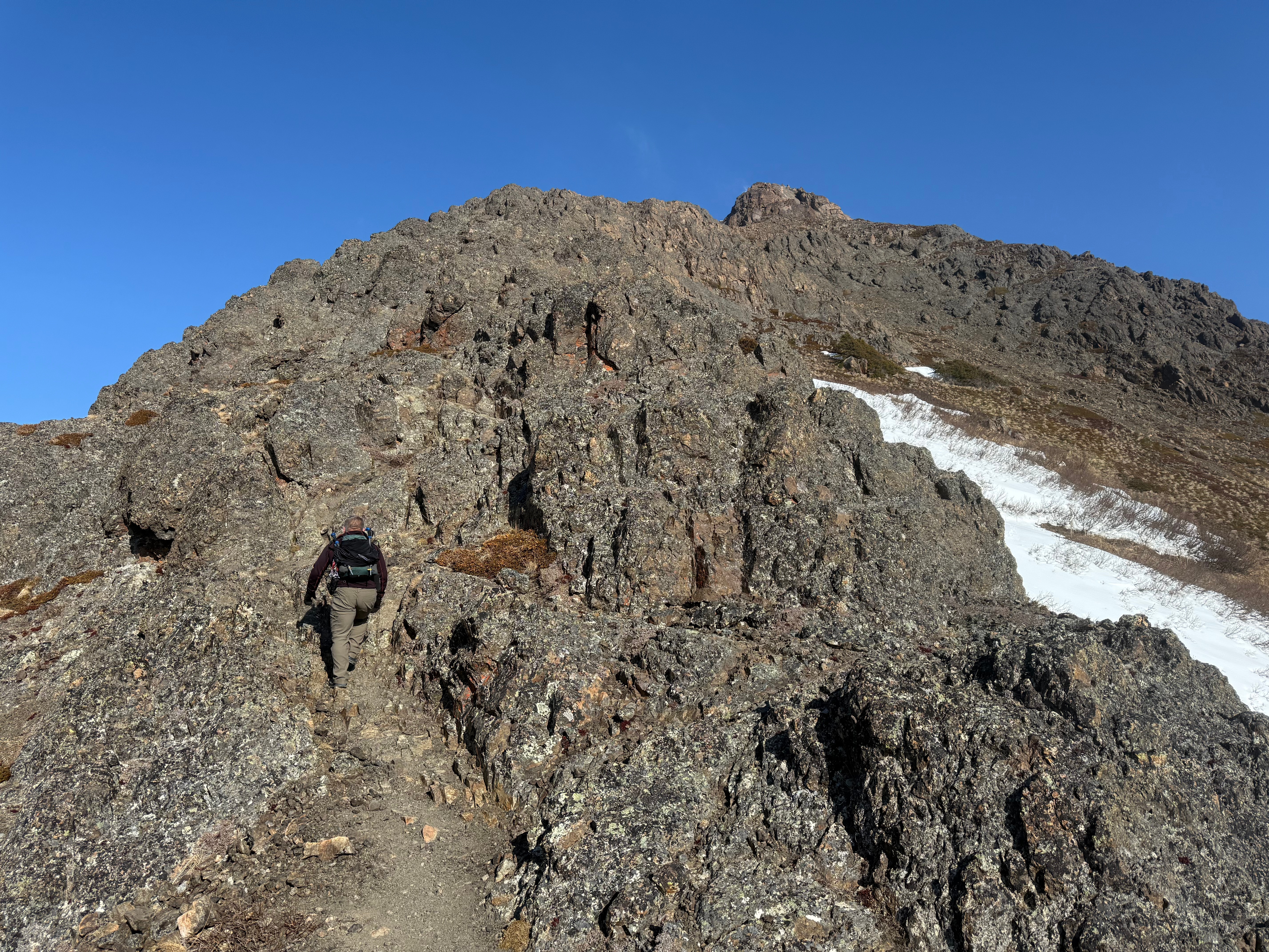 A hiker climbs up a steep, rocky trail. The summit is above and has snow blowing off of the high point.