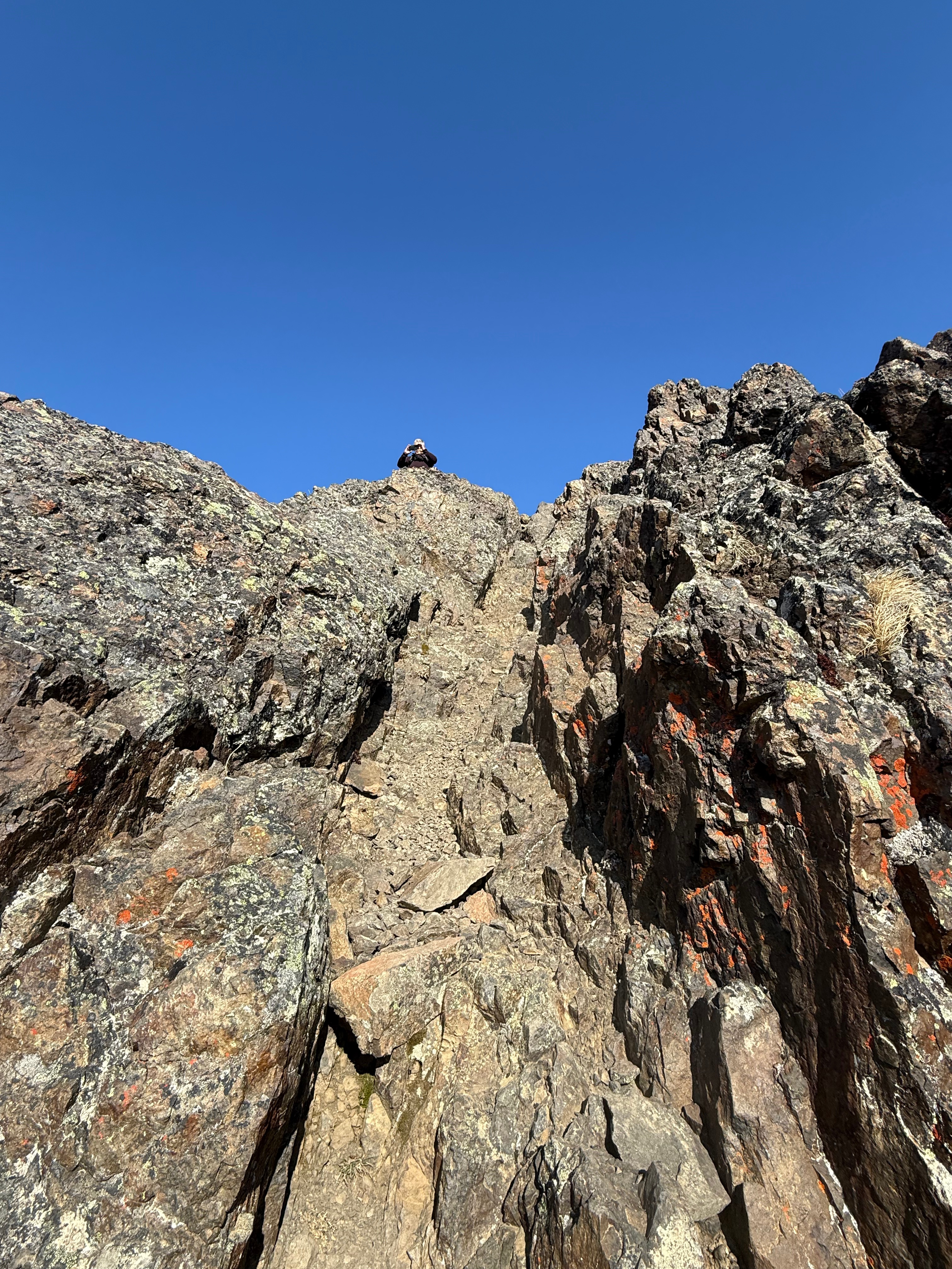 A hiker stands atop the vertical rock face, looking down to take a photograph.