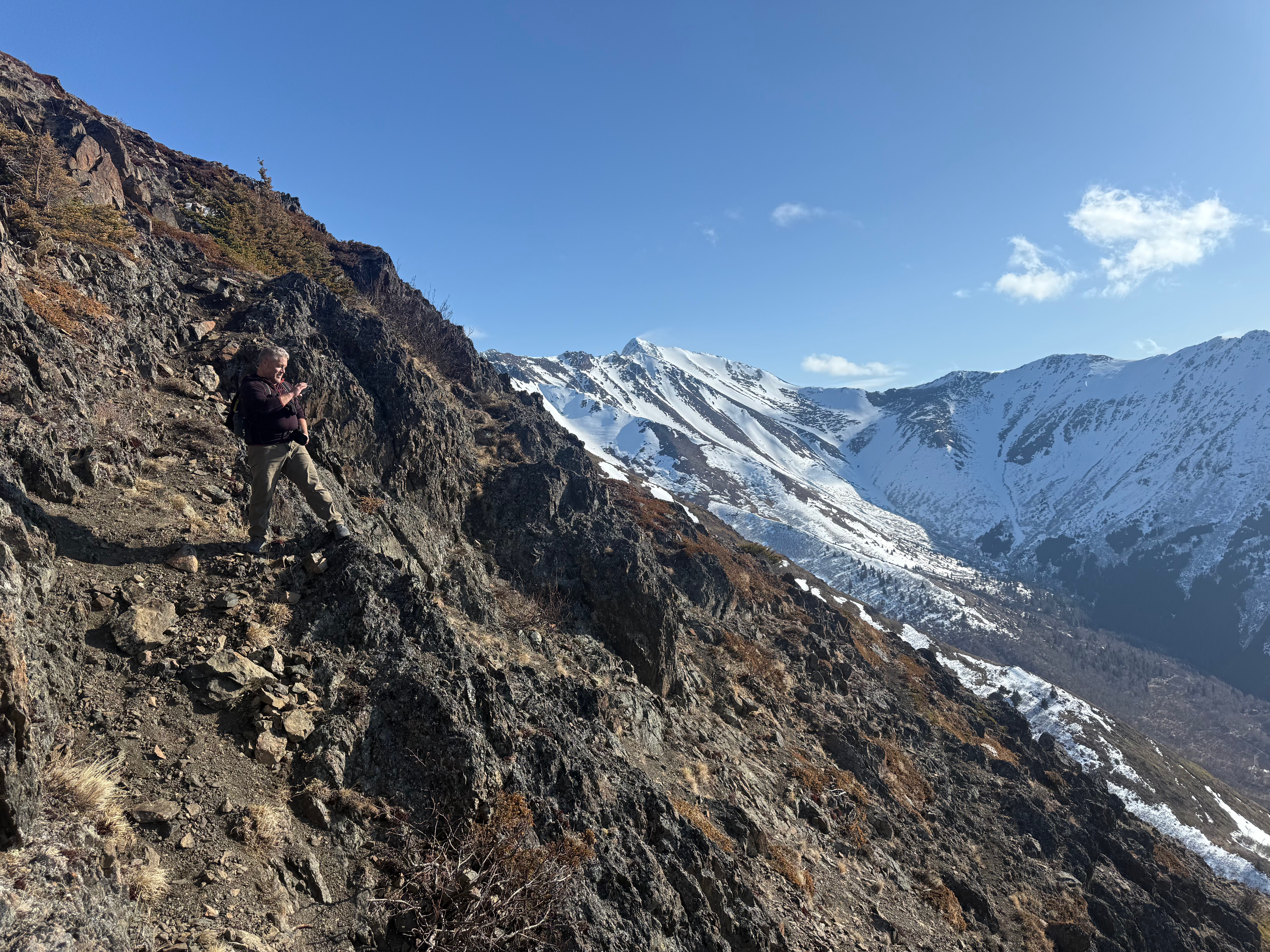 A hiker stands on a narrow, rocky trail that drops off steeply to the right. Snowcapped mountains are visible in the distance. Snow is blowing off the summit of a peak far in the distance.