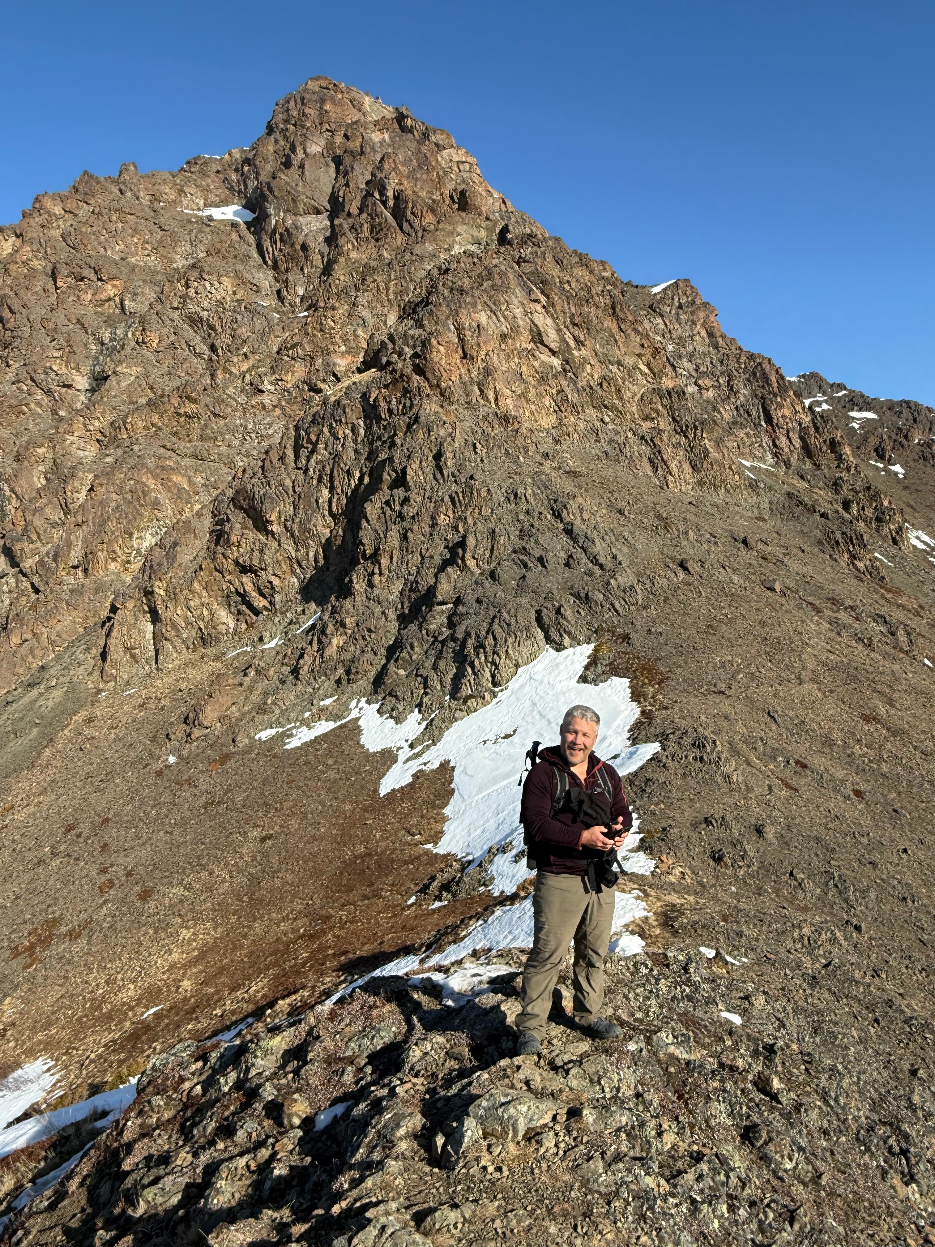 A hiker stands and smiles, the summit rising above in the background.
