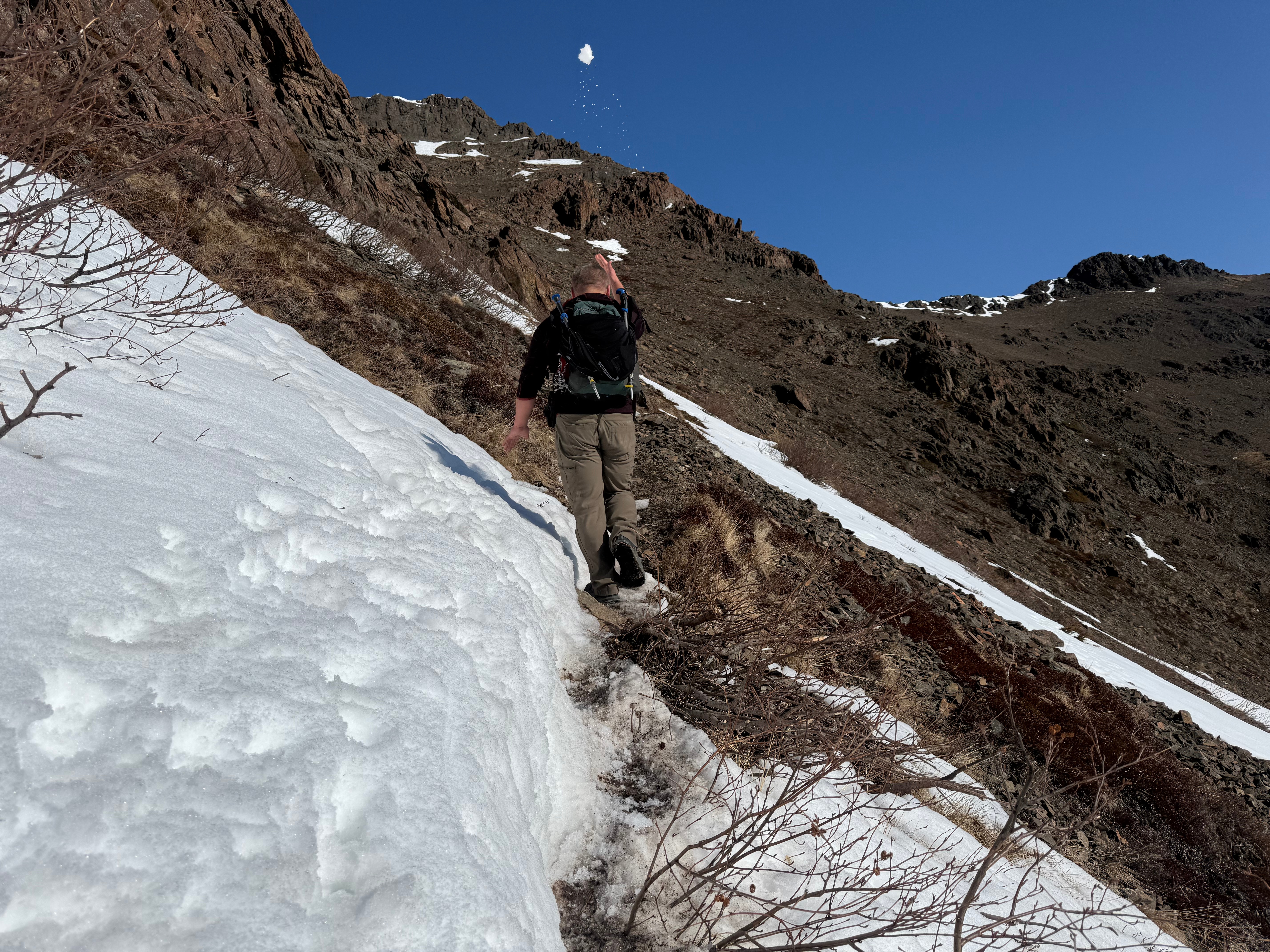 A hiker walks along a snowy slope that drops steeply off to the right. He is throwing a snowball back over his head towards the photographer.