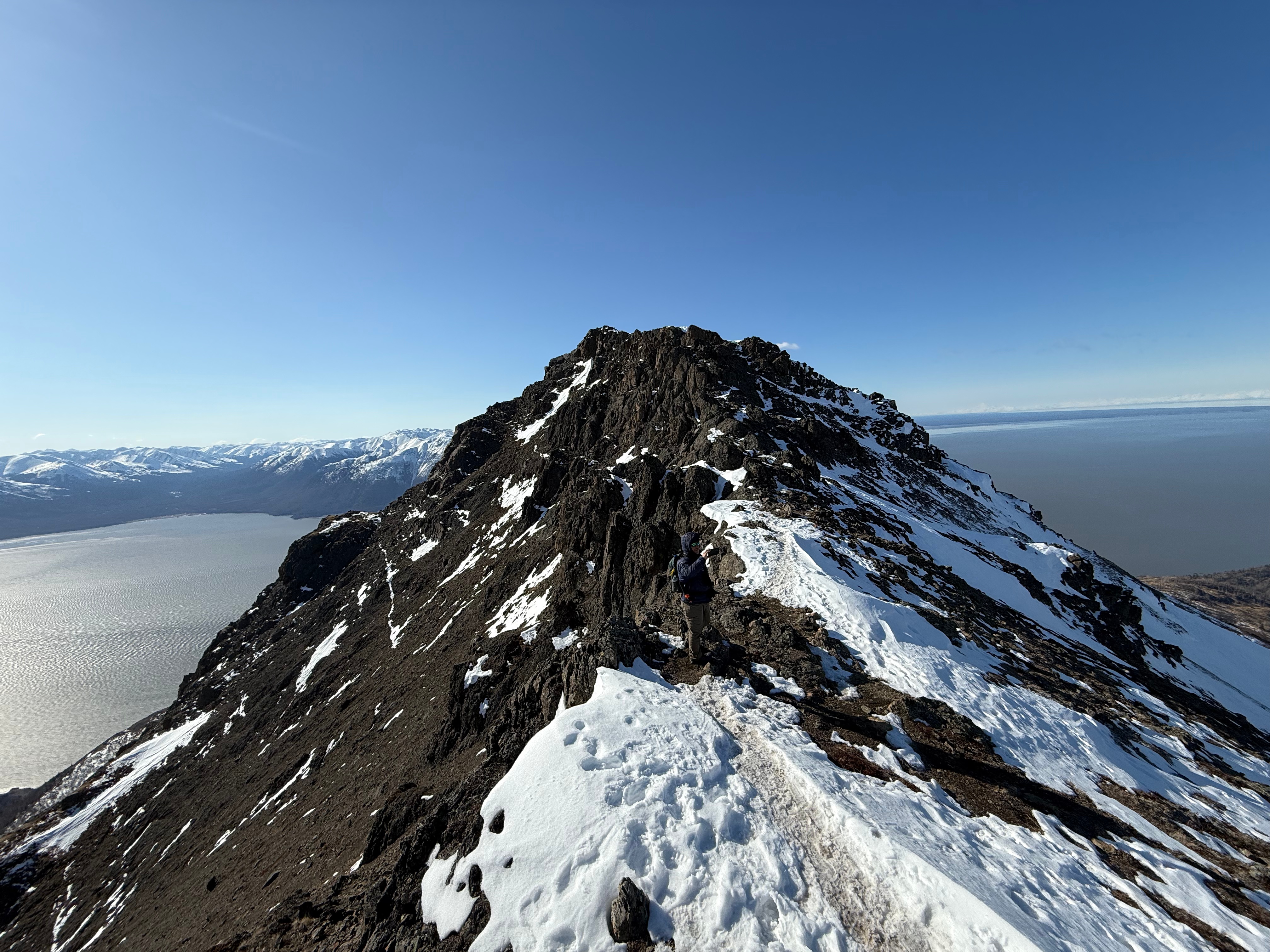 A hiker takes a photo while standing on the summit ridge. The summit is not far. Snow covers the shaded slopes on the right side of the photo.
