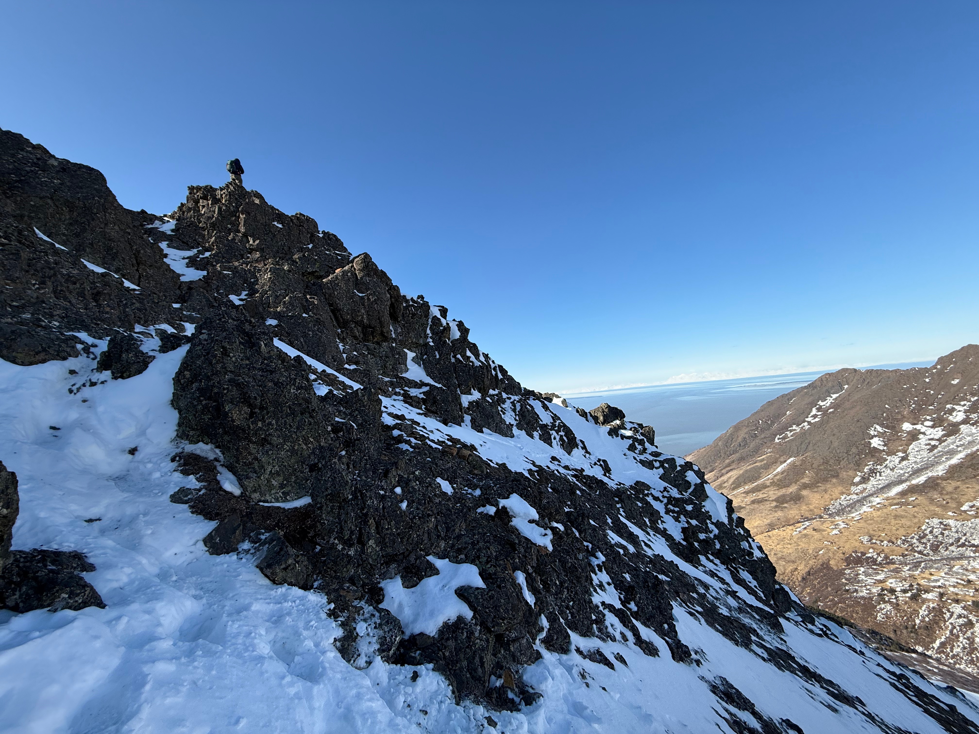 A hiker stands high atop a rock. The steep, shaded slopes are covered in snow and fall away to a sunny valley.