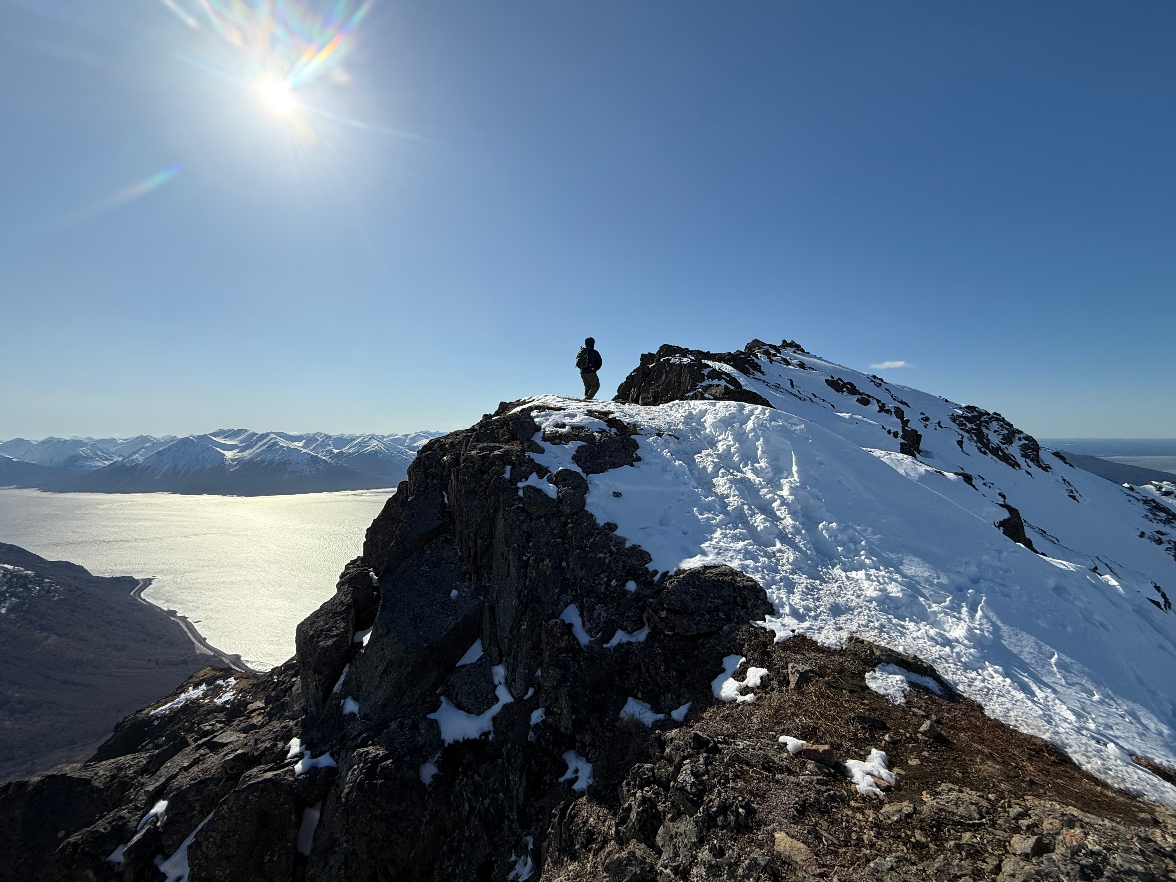 A hiker stands atop a point just below the summit. The ocean and highway are far below.