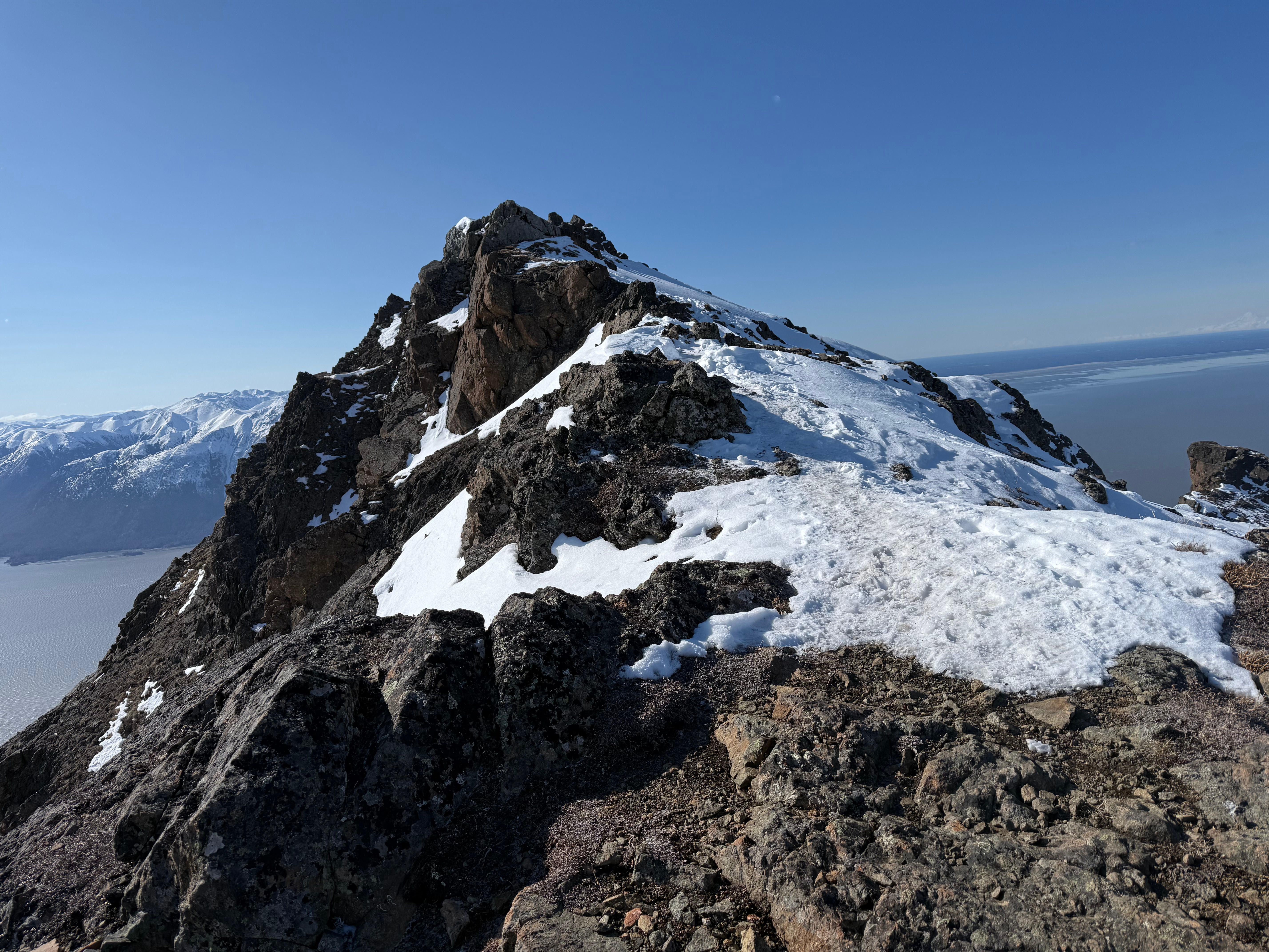 A snowy path leads the last short distance to the summit. The ocean is far below. A volcano is far in the distance across the ocean.