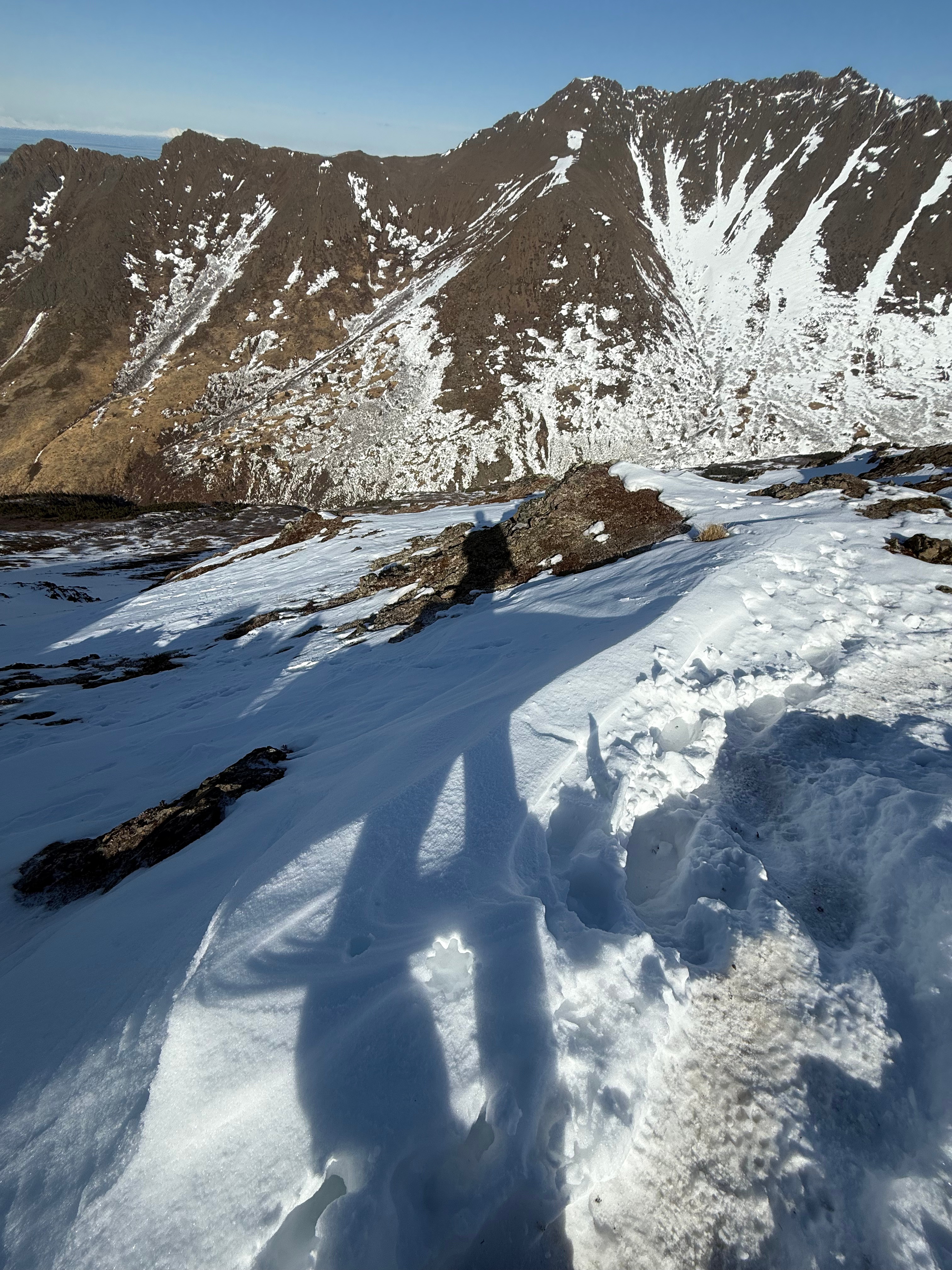 A long shadow of the photographer stretches down the sunny, snow covered slopes. Another mountain rises up in the background.