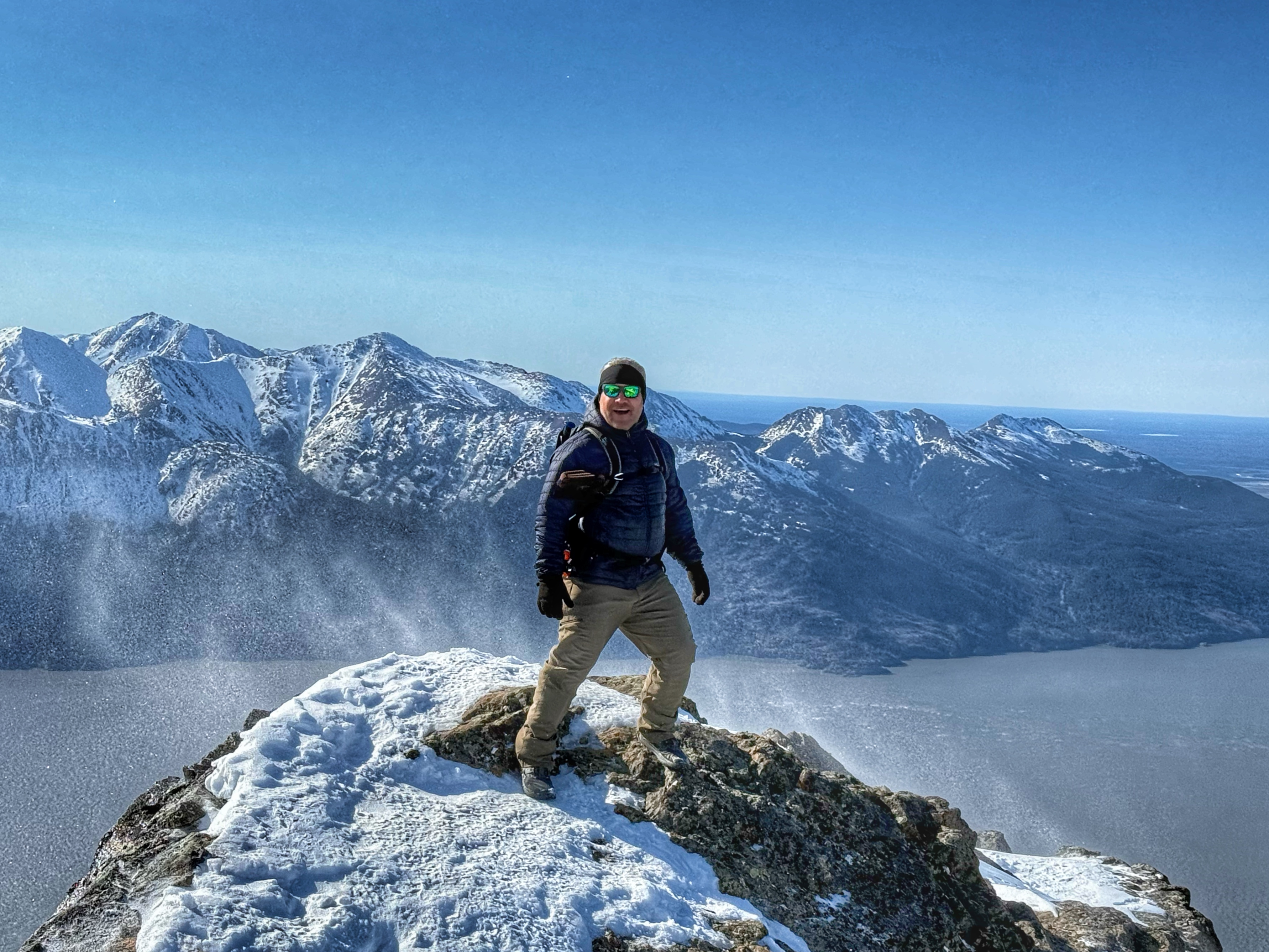 A person stands on top of a mountain summit, the ocean below and a mountain range on the other side of the inlet. Snow swirls around the hiker.