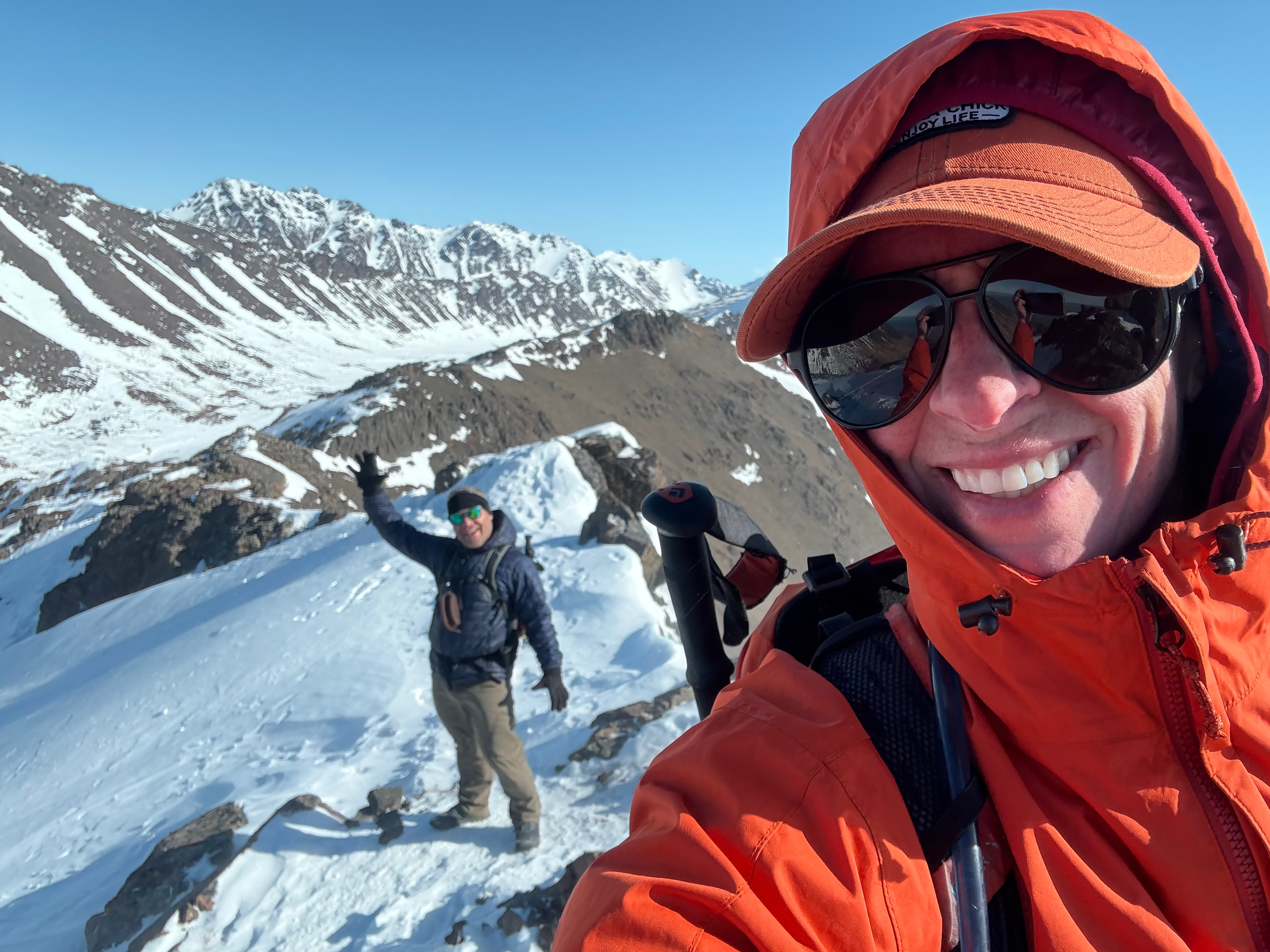 A selfie of the author with her friend in the background waving. Standing on the snow covered ridge. Mountains rise up in the distance.