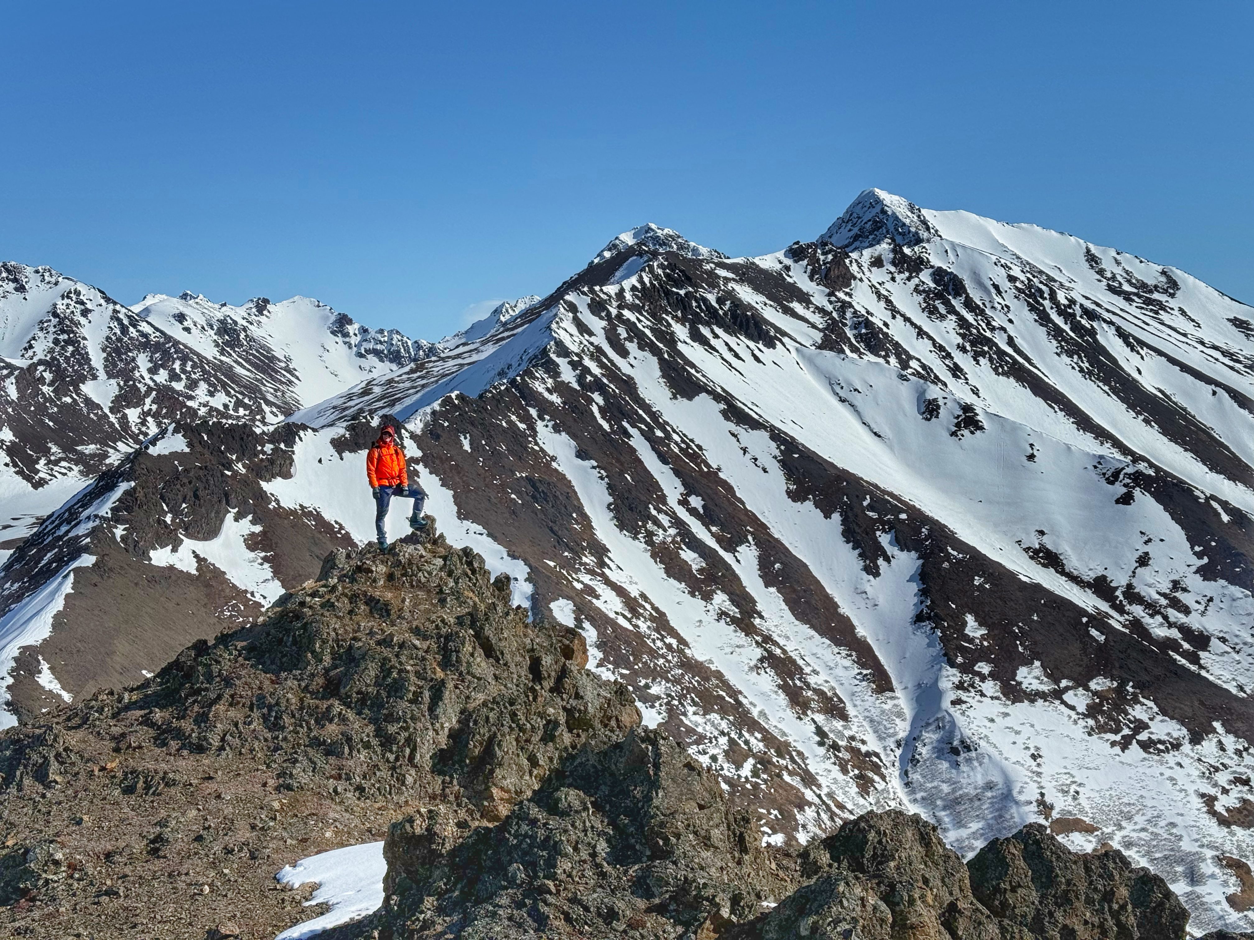 A person stands on a high rock in front of a tall mountain ridge.