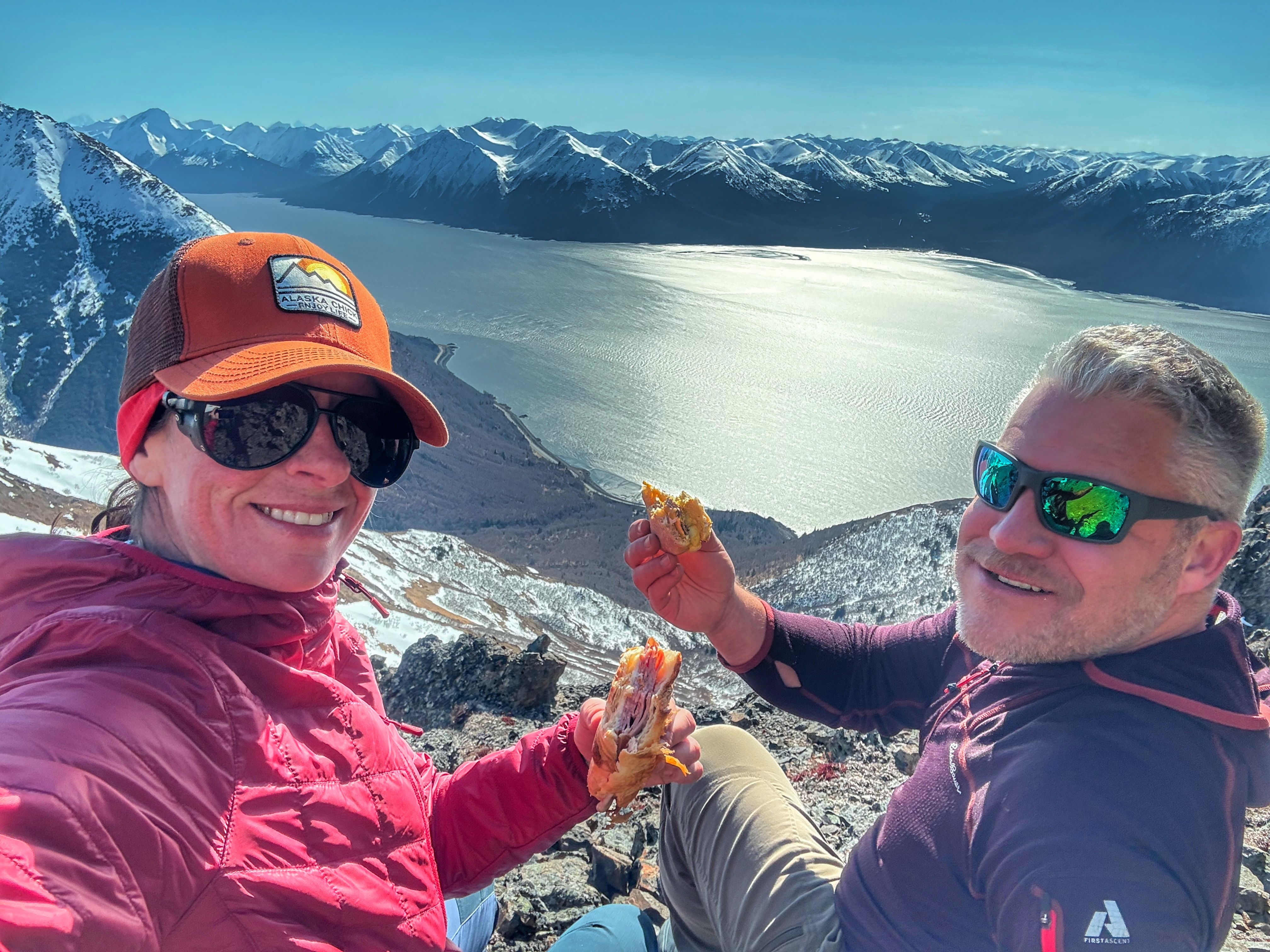 The author and her friend are sitting on top of a steep slope holding up sandwiches and smiling. The ocean is below and mountains rise up across the inlet.