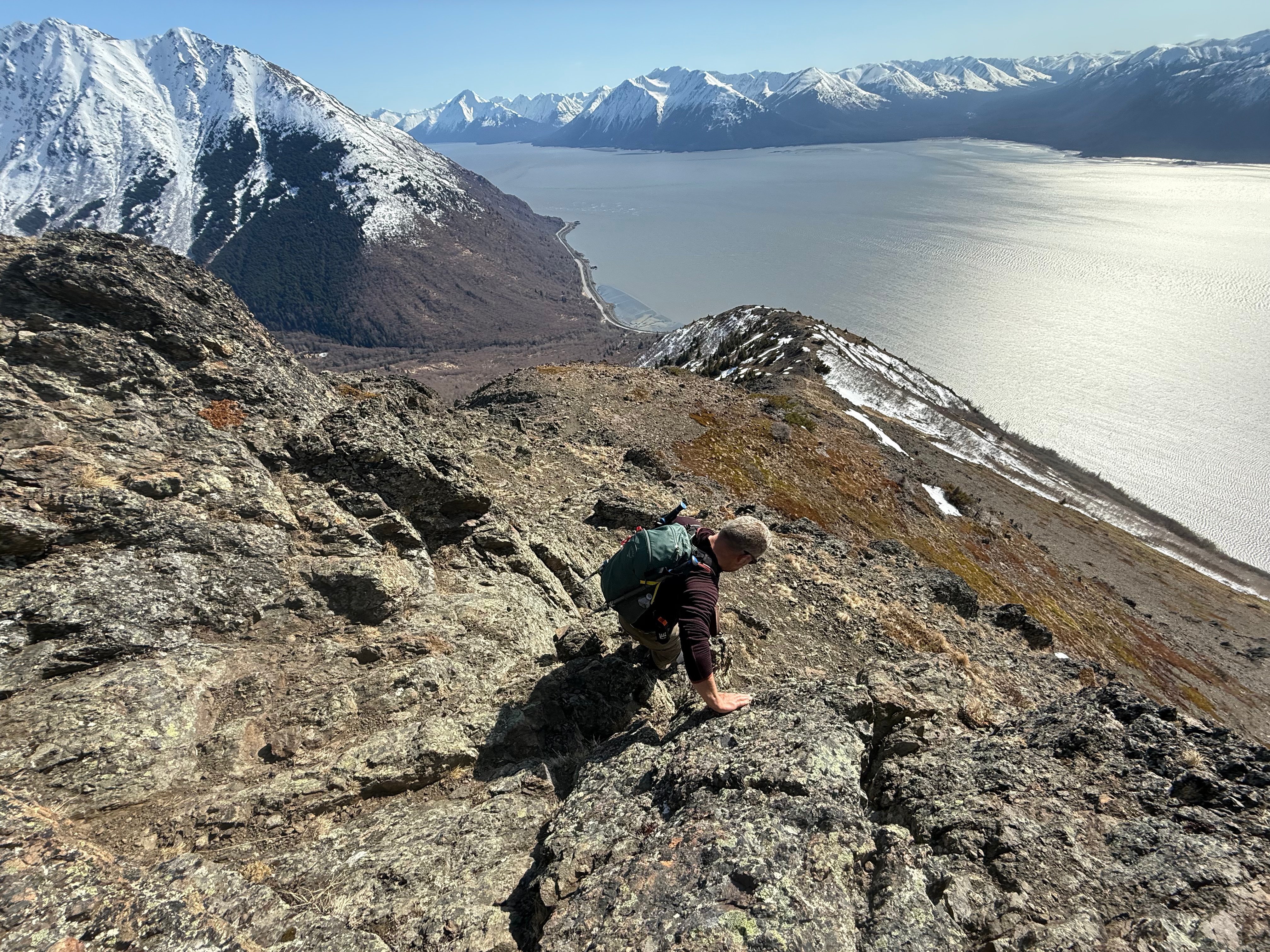 A hiker uses his hands to balance while descending a steep section of trail. The ocean is below.