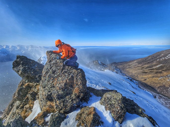 A person in a bright orange jacket clings to a rock on the summit of a mountain, battered by high winds